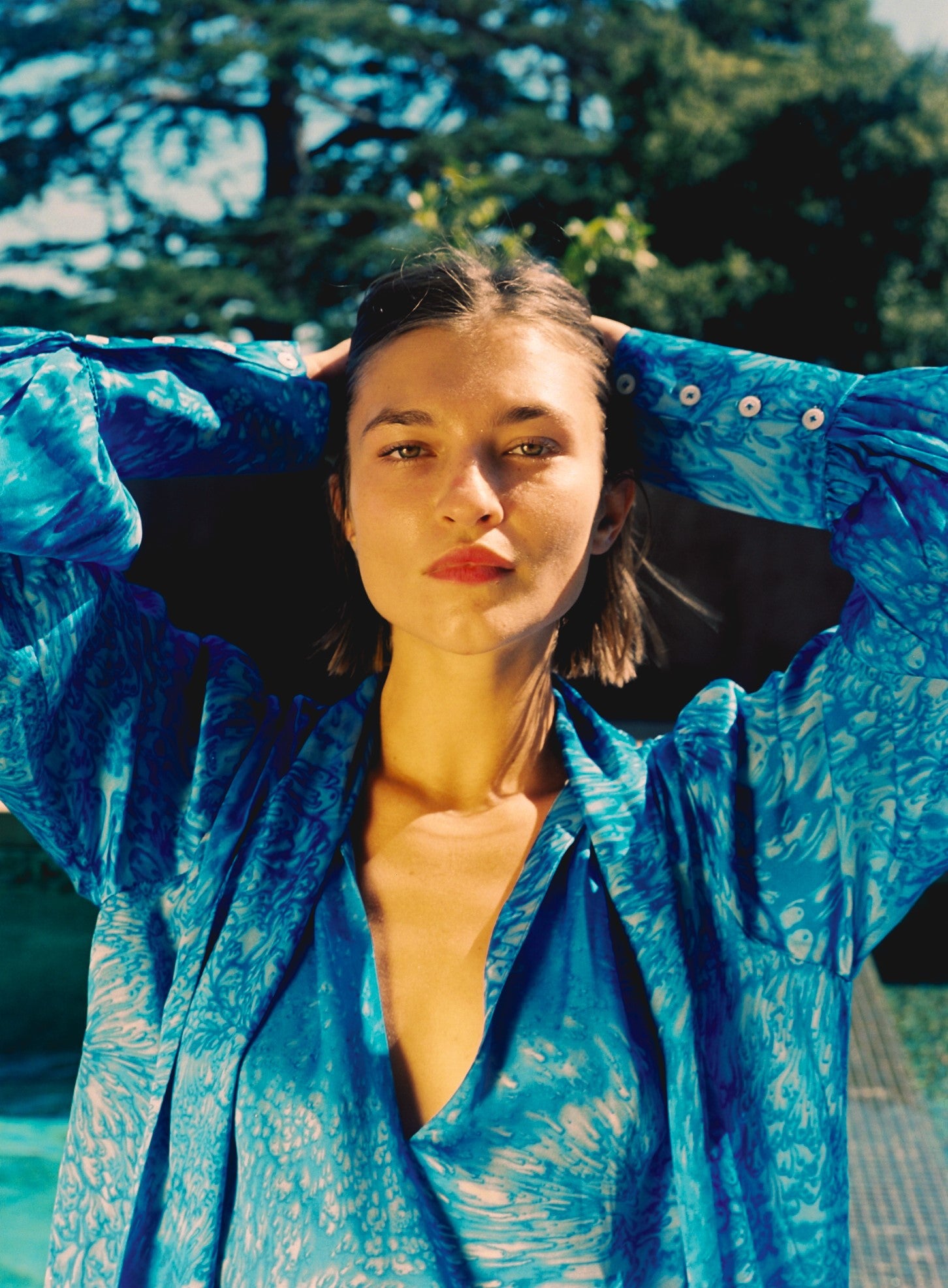 A woman is standing outdoors with arms raised, wearing a Catherine Gee's Aussie Short Dress - Blue Dye. The dress has a loose fit with a deep V-neckline and button details on the sleeves.  The background features greenery and a blurred tree, suggesting a natural setting that highlights the vibrant color and pattern of the dress, making it visually appealing and relevant for fashion or clothing discussions.
