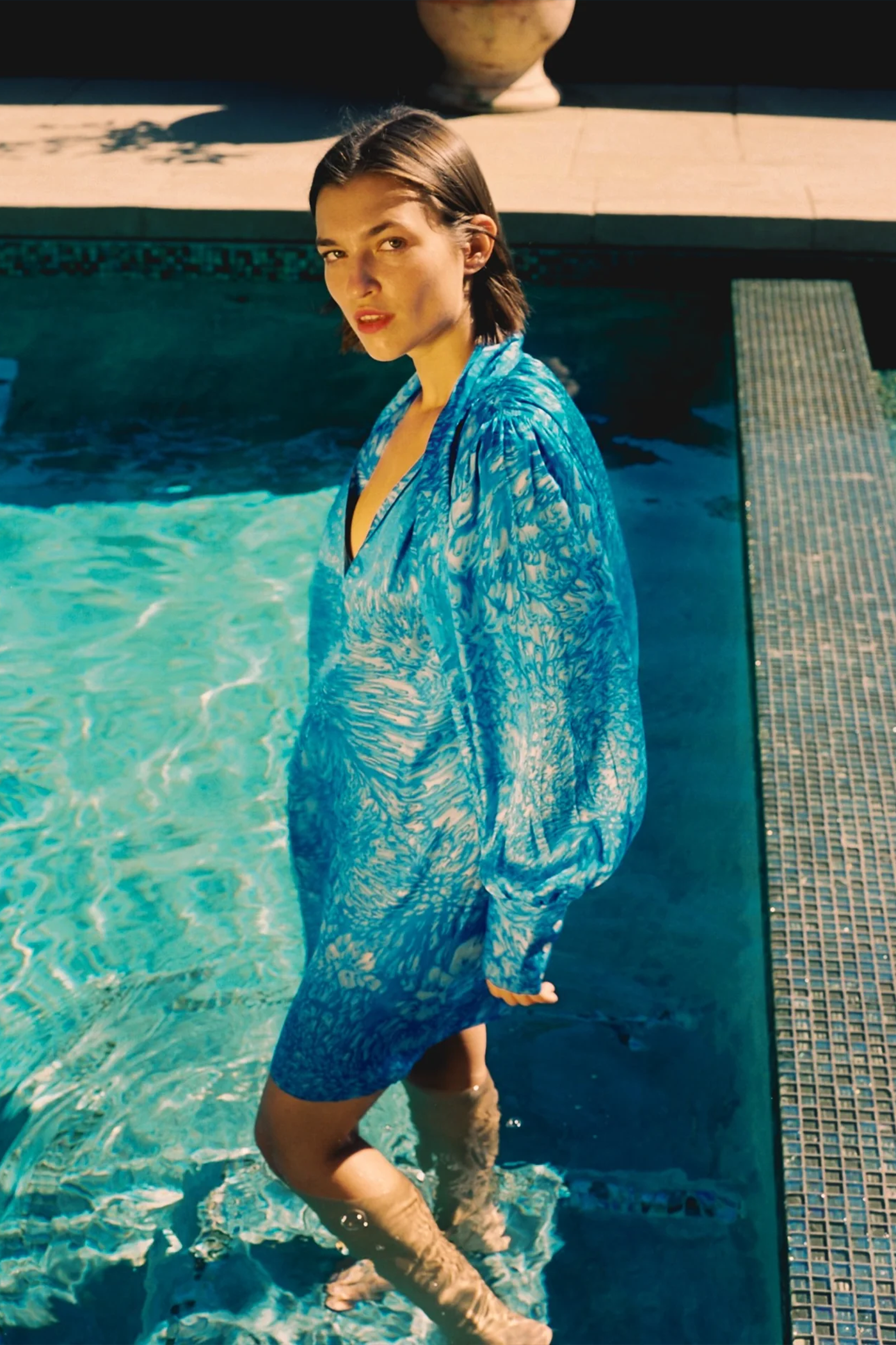 A woman is standing in a pool partially submerged in water wearing Catherine Gee's Aussie Short Dress - Blue Dye with the lower part of her dress and legs visible in the pool. The background includes the edge of the pool and a potted plant highlighting the dress design and how it looks when wet, showcasing its appearance and texture in a unique setting.