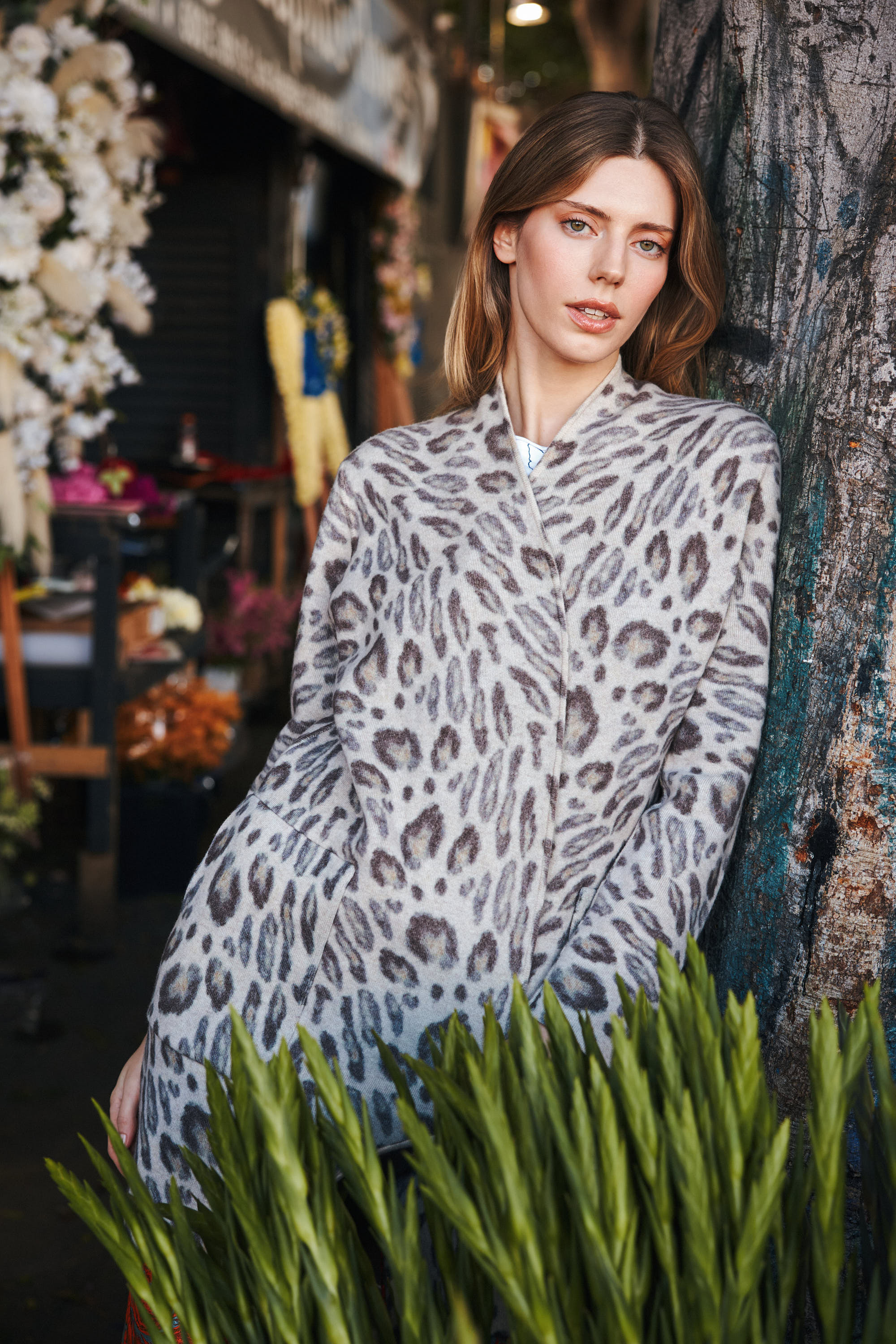 A woman standing and leaning against a tree, wearing a Catherine Gee Double Face Cashmere Reversible New Leopard coat features a sophisticated leopard print pattern, adding a touch of luxury and style. The elegant and versatile design of the coat makes it a standout piece, perfect for making a fashionable statement.