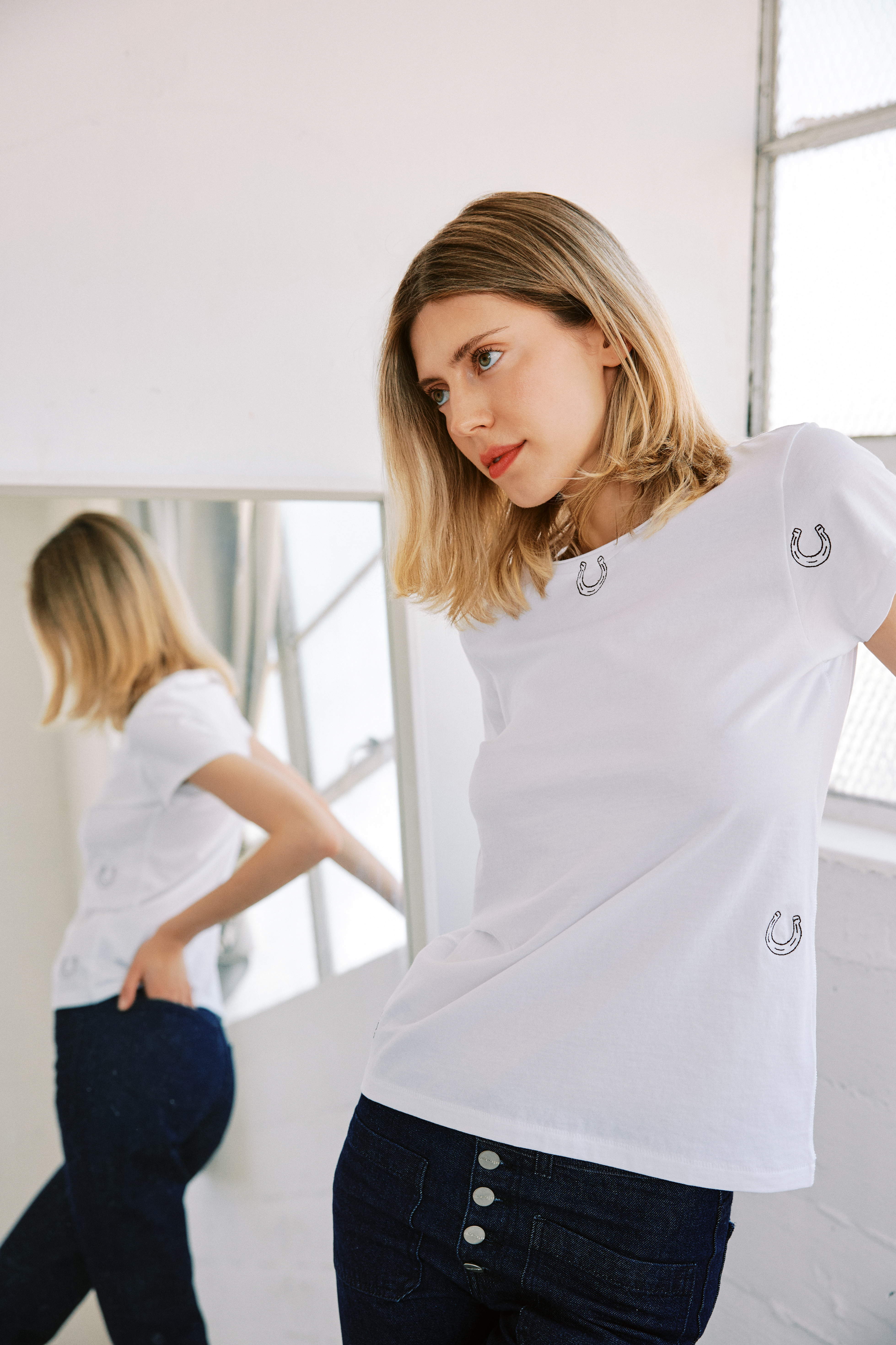 Catherine Gee Embroidered Cotton Horseshoe white t-shirt features small embroidered horseshoe designs scattered across the chest and near the waist. She is standing in front of a mirror, providing a view of both the front and back of the t-shirt paired with dark blue jeans with multiple buttons. The natural light filters through a window, highlighting the t-shirt's casual elegance and unique design.