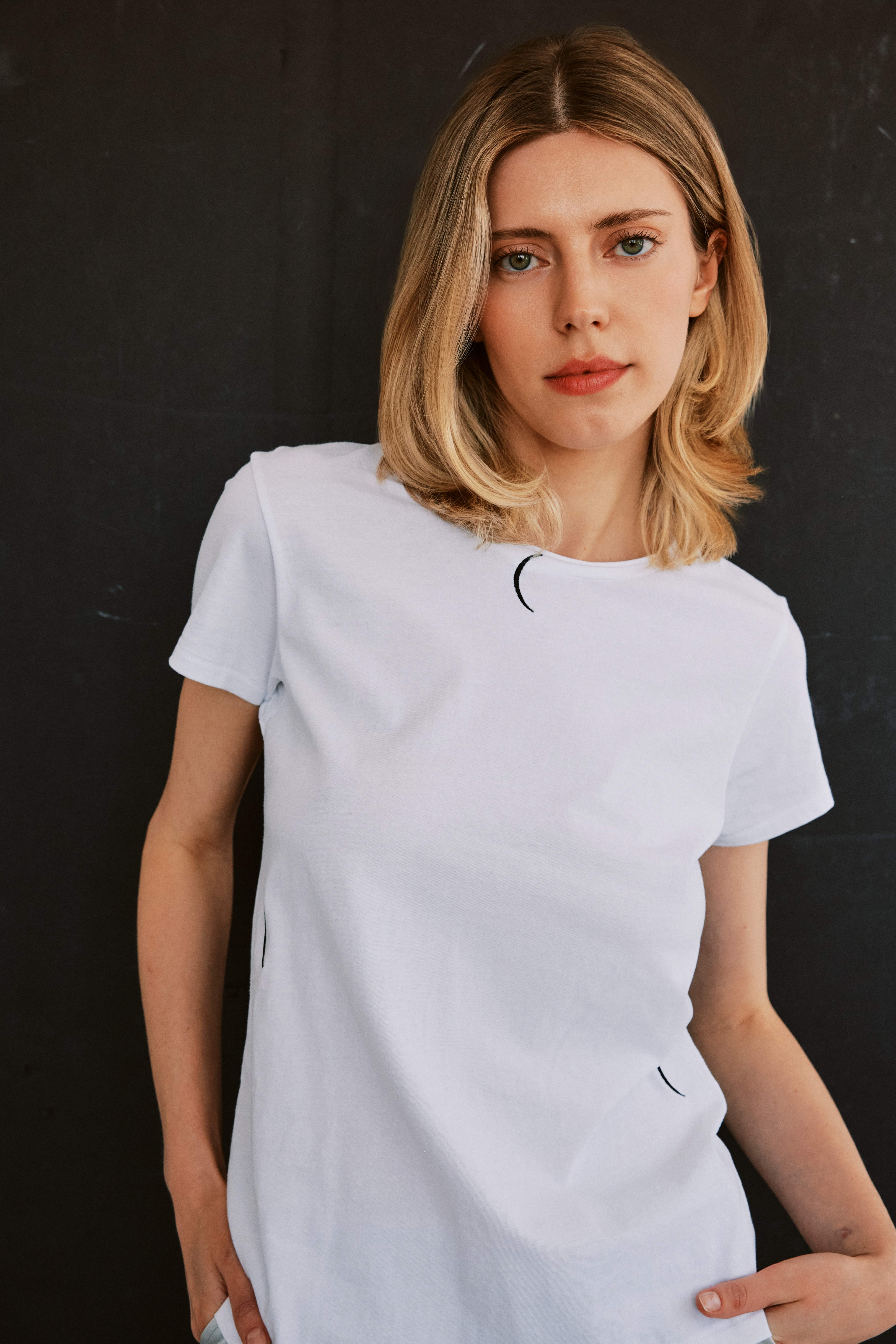A woman stands against a dark background, with one hand in a pocket and the other resting on the hip wearing a white Catherine Gee Embroidered Cotton Crescent T-shirt features small, black crescent moon embroidery in various places, including near the neckline, on the side, and near the bottom hem. This relaxed-fit t-shirt offers a simple yet stylish design, perfect for casual wear with a touch of mystique.