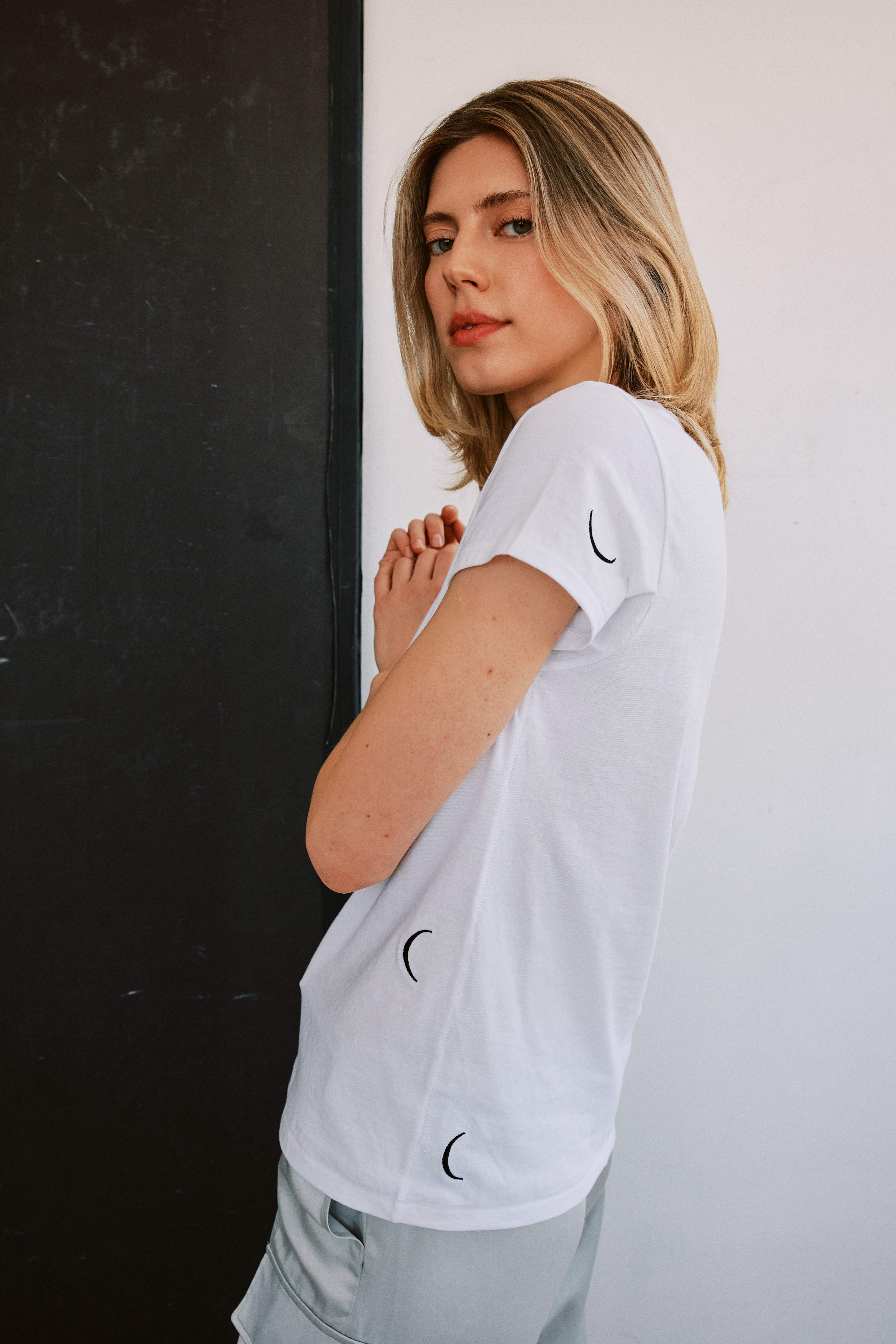 A woman standing sideways, with the left arm bent and the hand near the face wearing a white Catherine Gee Embroidered Cotton Crescent t-shirt features small crescent moon embroidery designs on the sleeve and side on a black and white wall background, highlights the minimalist and elegant design of the t-shirt, making it interesting for those who appreciate unique fashion items.
