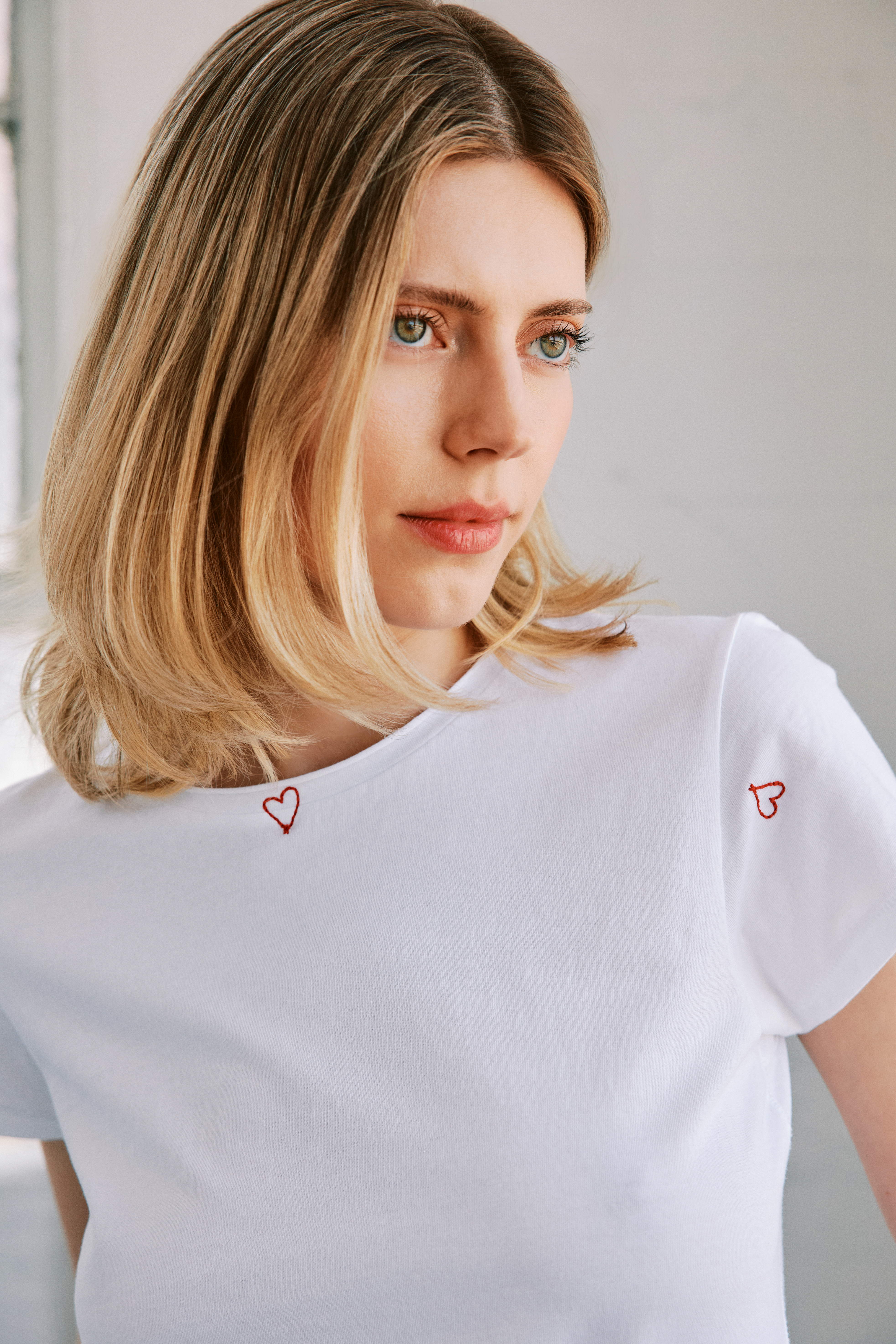 A close-up of a woman wearing a Catherine Gee Embroidered Cotton Red Heart white t-shirt features small red heart embroidery designs, with one on the left shoulder and another near the right sleeve. She has shoulder-length blonde hair, creating a simple yet elegant look. The delicate heart embroidery adds a charming touch, making this t-shirt a stylish and versatile piece for casual wear.