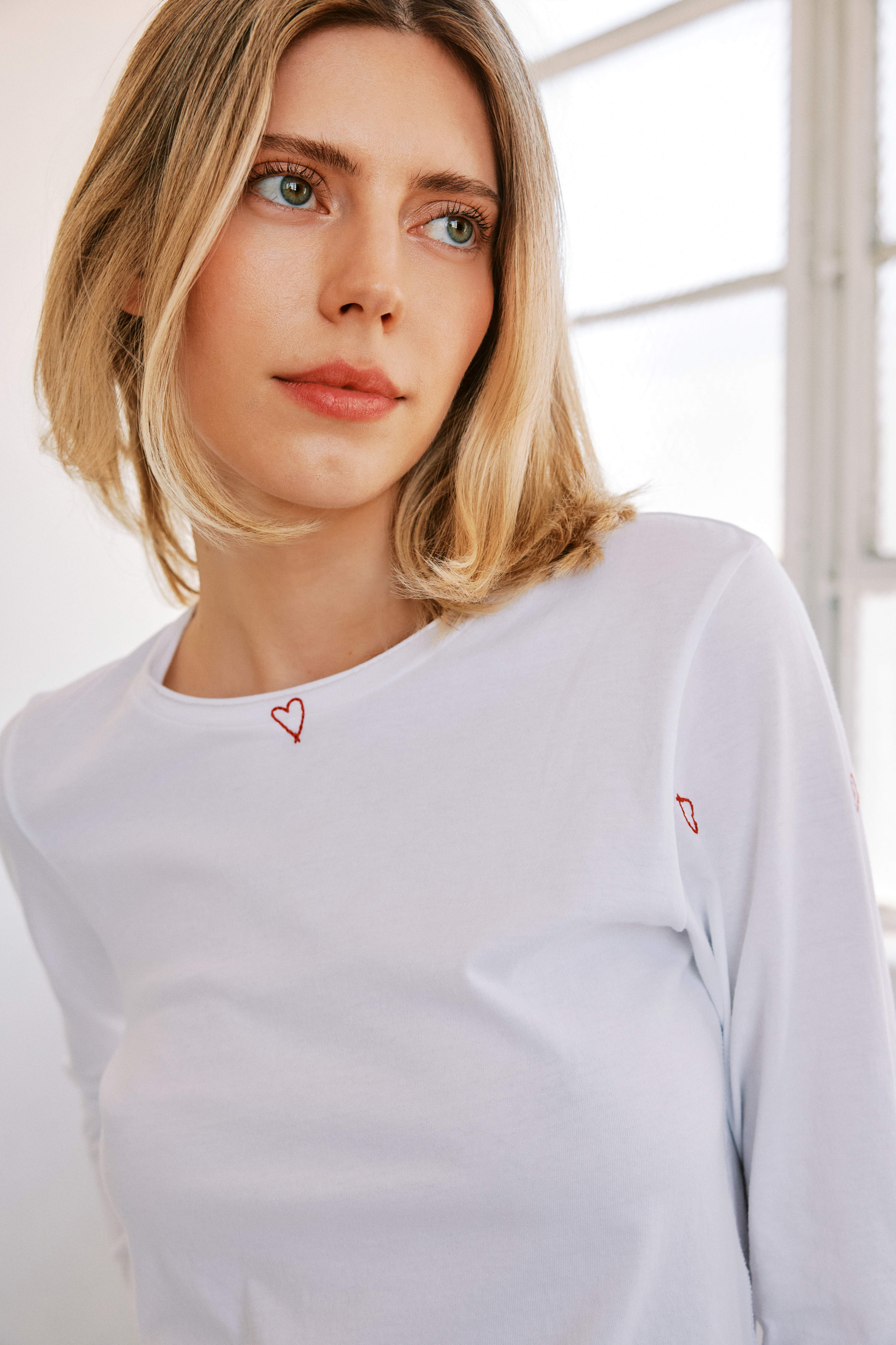 A woman wearing a Catherine Gee Embroidered white Cotton Long Sleeve Red Heart T-Shirt with small red heart embroidery details on the chest and sleeve. The background appears to be a bright room with large windows highlighting the simple yet elegant design of the shirt.