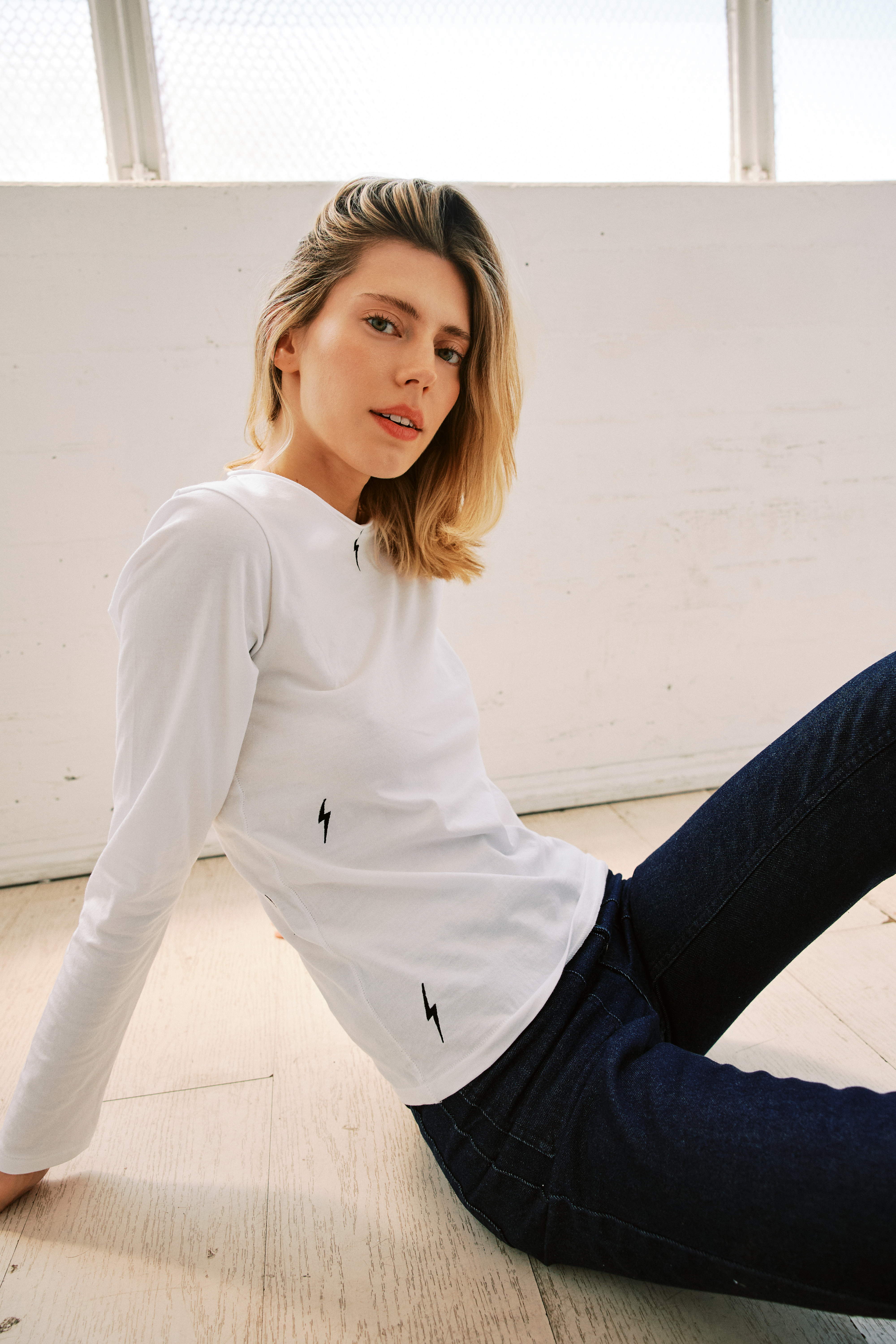 A woman is seated on the floor in a light-colored room with large windows. She leans back, supporting herself with one arm wearing a Catherine Gee Embroidered Cotton White Long Sleeve Bolt T-Shirt, which features small black embroidered lightning bolt designs on both the front and the sleeves. The shirt's simple yet bold embroidery makes it a stylish and comfortable choice for everyday wear.