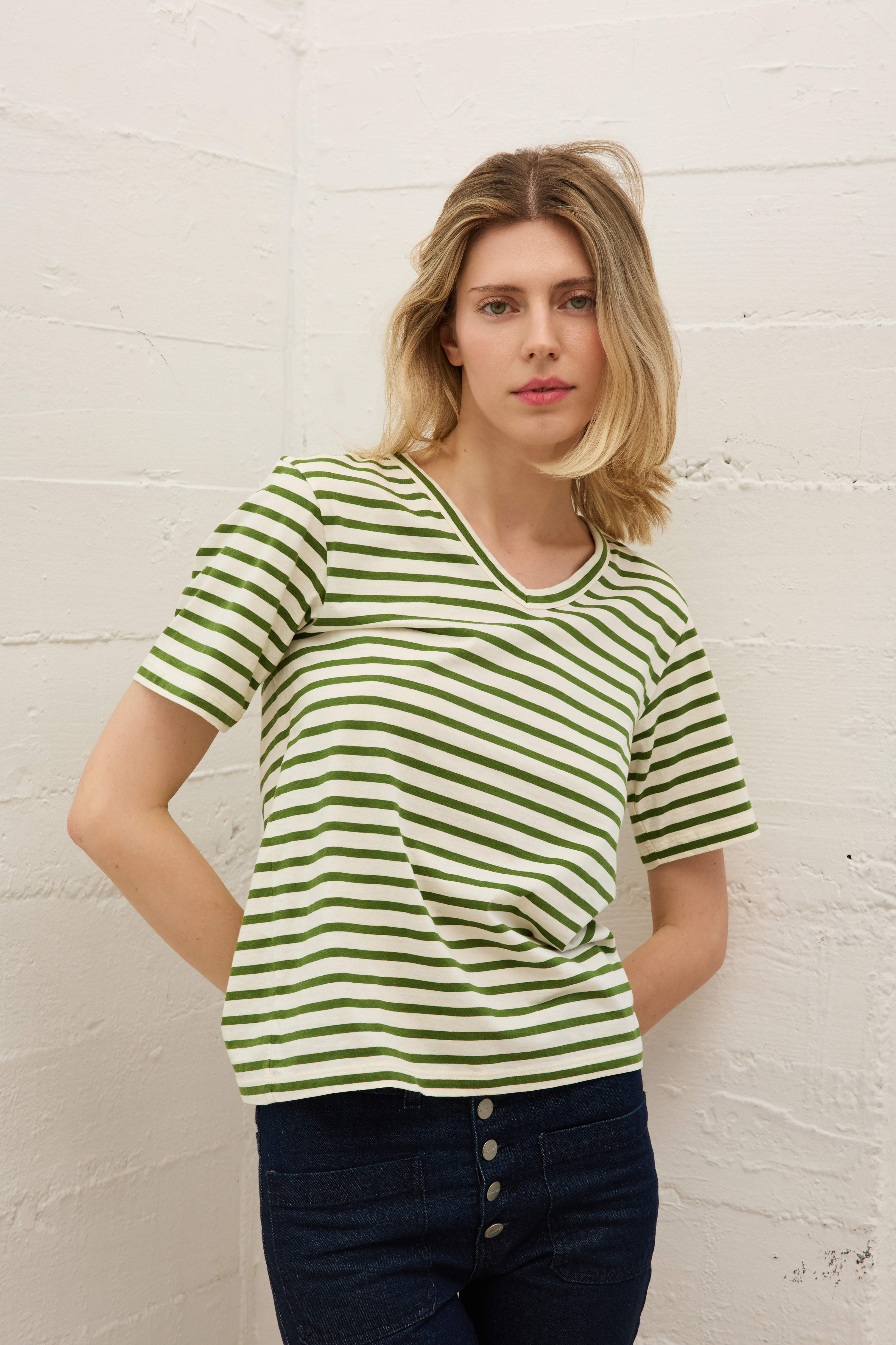 A woman wearing the Catherine Gee Jen Relaxed Shortsleeve - Green Stripe tee features horizontal green and white stripes and has a relaxed fit with short sleeves paired with dark blue high-waisted pants that have buttons visible at the front. The background is a plain, light-colored wall with a textured surface. The overall look is casual and stylish, perfect for a laid-back yet chic outfit.