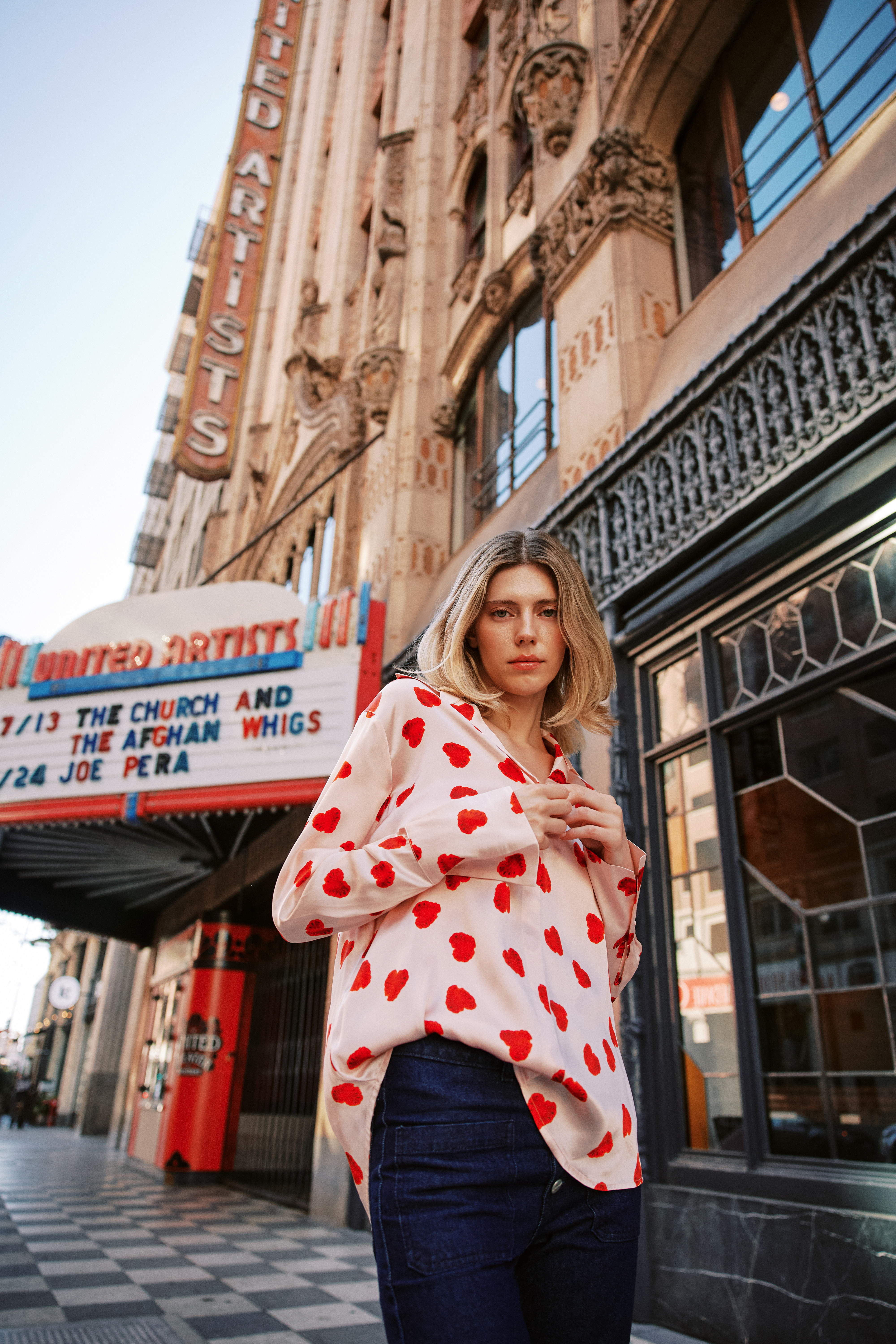 A woman standing in front of the United Artists Theatre, wearing a Catherine Gee Daria French Cuff Silk Raspberry Heart blouse. Features a white base color with a pattern of red hearts and a loose fit with French cuffs paired with dark blue jeans. The background includes the theatre's marquee, which lists upcoming shows, adding an interesting contrast to the modern fashion of the blouse.