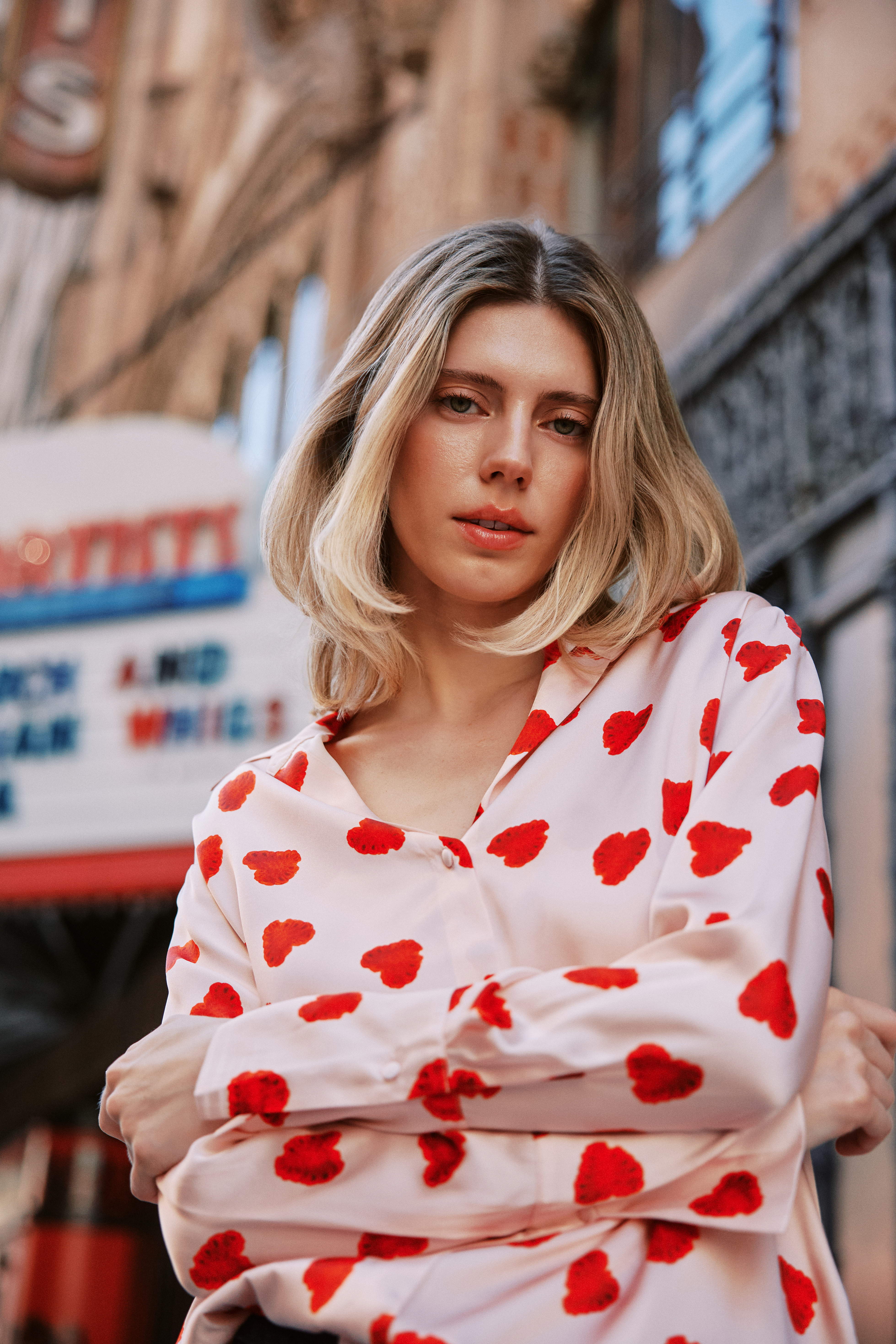 A woman wearing a Catherine Gee Daria French Cuff Silk Raspberry Heart blouse features a white base color with a pattern of red hearts, creating a playful and charming look. It has a classic collar, long sleeves with French cuffs, and a relaxed fit. She is standing with arms crossed in an urban setting, with a blurred sign and building in the background, highlighting the blouse's eye-catching and stylish design.