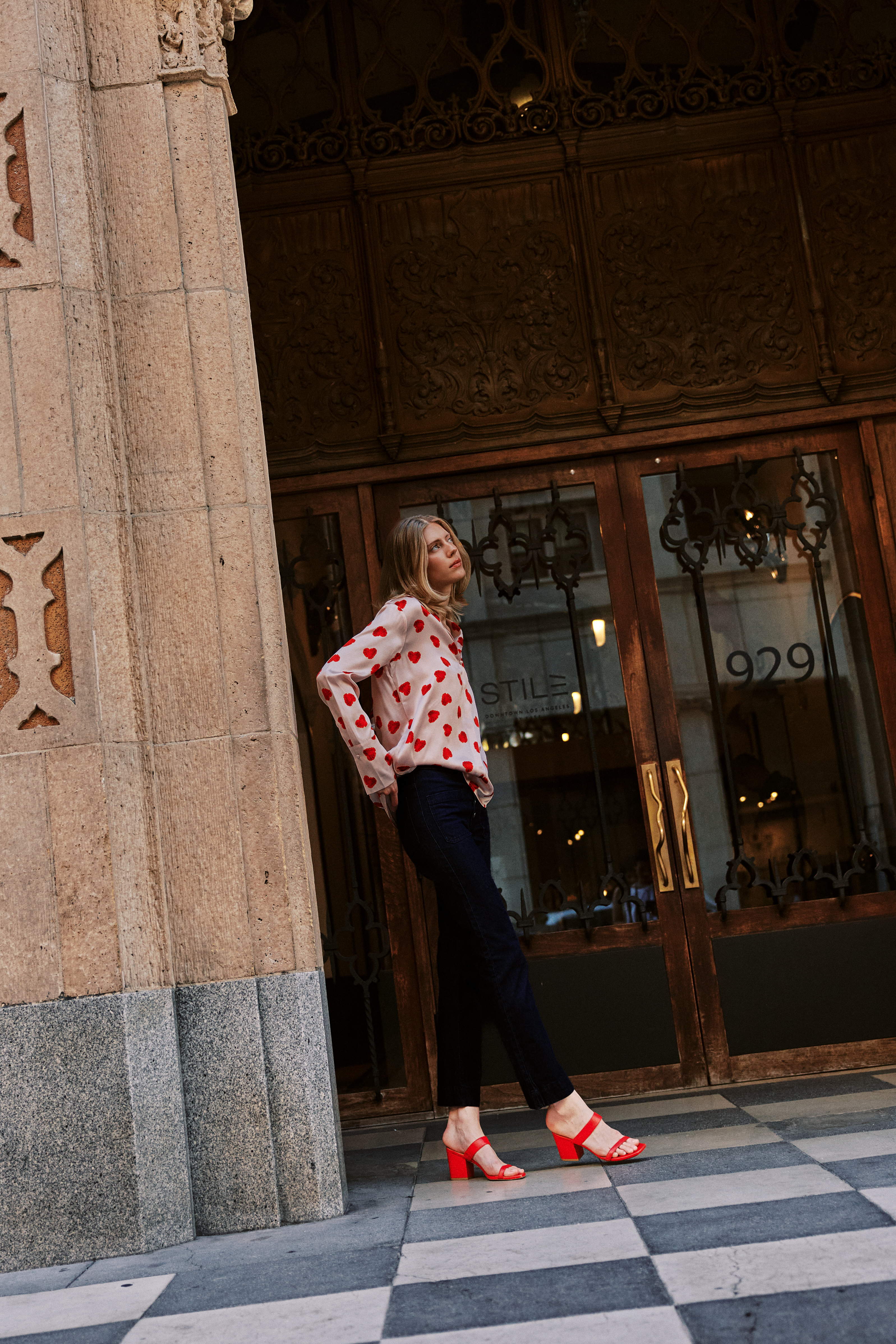 A woman standing in front of a building with ornate wooden doors and a stone facade wearing a Catherine Gee Daria French Cuff Silk Raspberry Heart Blouse, which features a pattern of red hearts on a white base color paired with dark pants and red high-heeled sandals. The setting appears to be urban, with a checkered floor pattern adding to the aesthetic appeal.