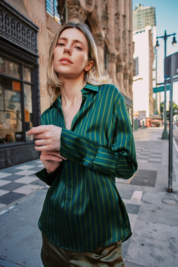 A woman stands on a city sidewalk in front of a building with ornate architecture and a checkered sidewalk pattern wearing a Catherine Gee Daria French Cuff Silk-Green Stripes blouse that features French cuffs and a relaxed fit.