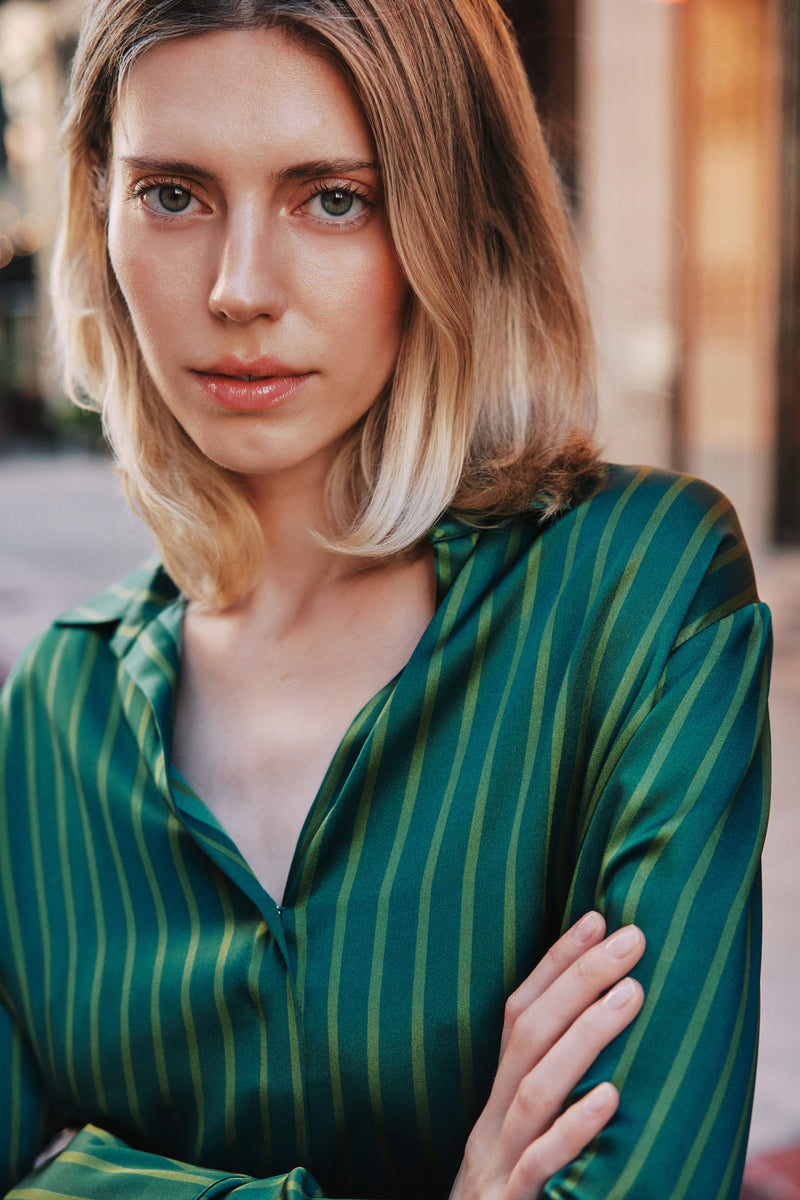 A woman with blonde hair standing with arms crossed wearing a Catherine Gee Daria French Cuff Silk-Green Stripes blouse features a deep V-neck and long sleeves.