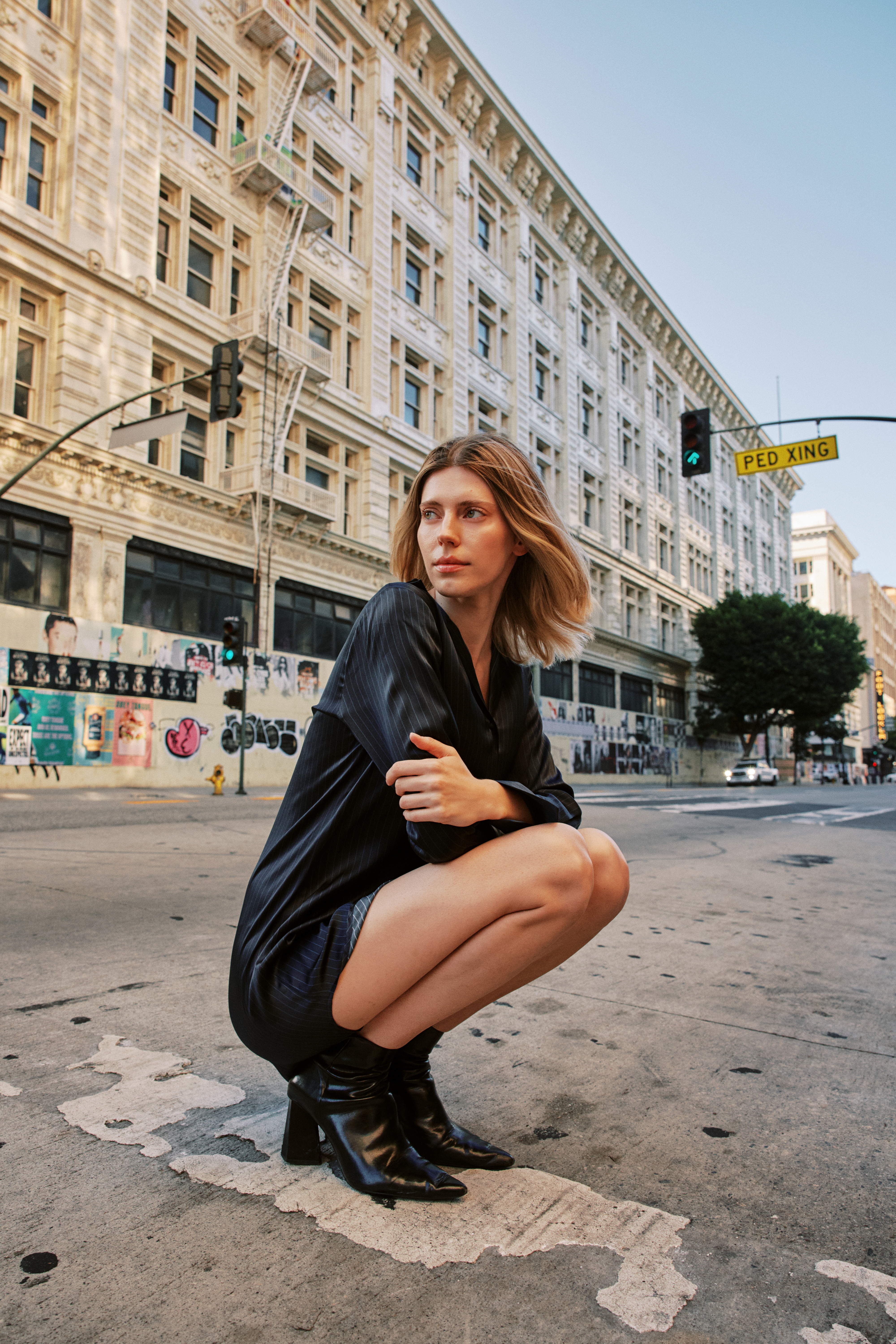 A woman crouching on a city street, wearing a Catherine Gee navy pinstripe Daria Dress and black ankle boots. The background features a large, ornate building with multiple windows, a 'PED XING' street sign, and graffiti-covered walls, adding an urban and artistic vibe to the scene.