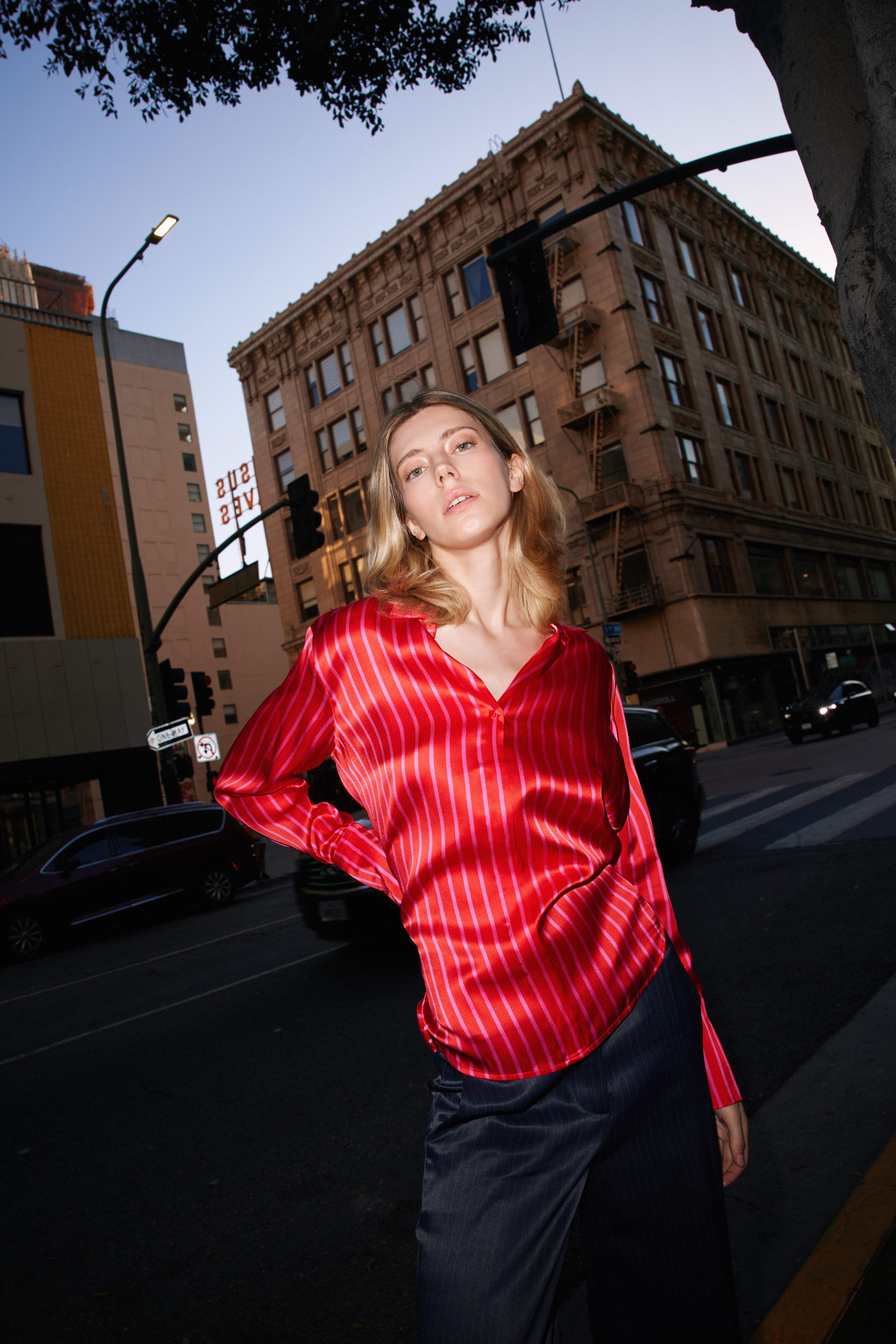 A woman standing on a city street, wearing a Catherine Gee Daria French Cuff Silk Red/Pink Stripe blouse features a deep V-neckline and long sleeves with French cuffs. The background includes an urban setting with tall buildings, streetlights, and cars, suggesting a busy city environment. The vibrant color and luxurious fabric of the blouse stand out against the urban backdrop, highlighting its stylish and elegant design.