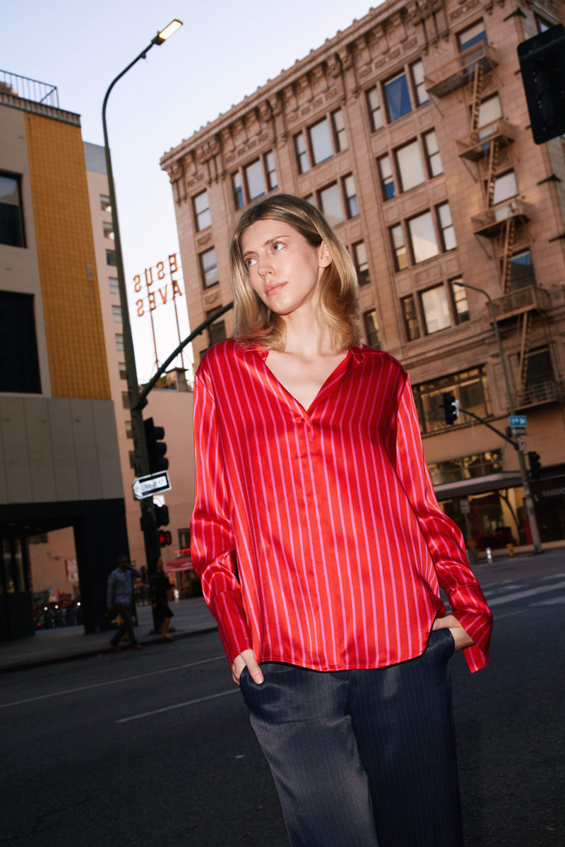 A woman standing on a city street with hands in her pockets, and the background includes urban buildings and a street sign wearing a Catherine Gee Daria French Cuff Silk Red/Pink Stripe blouse features a V-neckline and long sleeves with French cuffs. The vibrant color and elegant design of the blouse make it a standout piece in the urban setting.