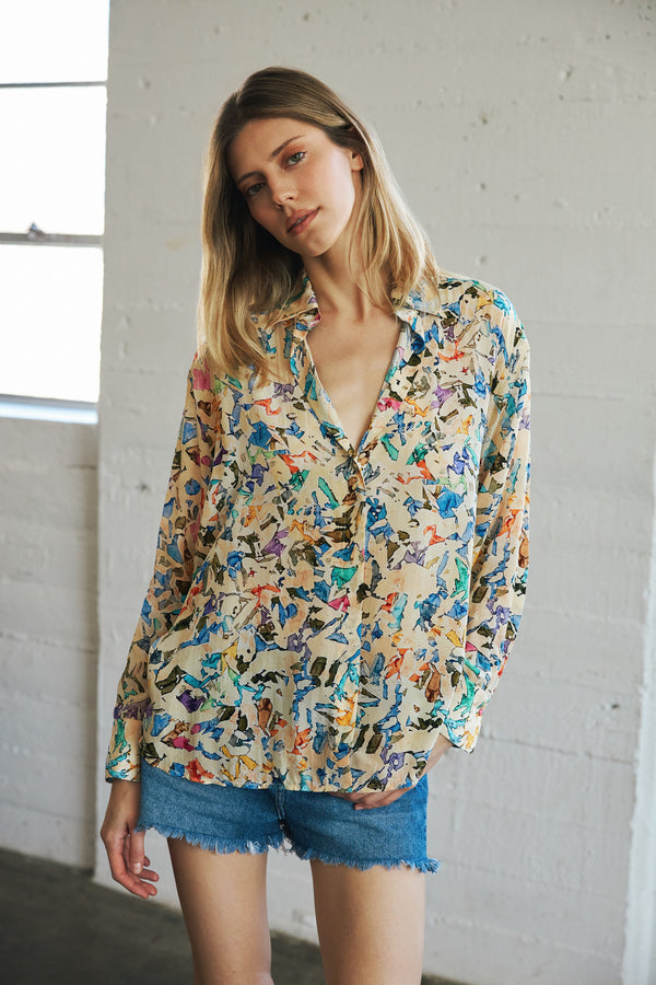 A woman wearing a Catherine Gee Dakota Blouse in the Museo print features a colorful, abstract pattern on a light beige background with a relaxed fit and long sleeves. It is paired with blue denim shorts with a frayed hem. The background shows a white brick wall and a window, emphasizing the blouse's unique design and casual style.