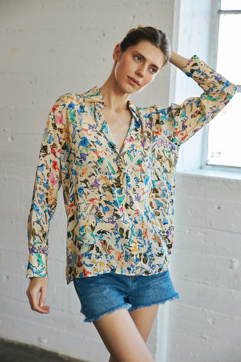 A woman is wearing a Catherine Gee Dakota Blouse in the Museo print, featuring a colorful abstract pattern. The blouse has a V-neckline and long sleeves. She is also wearing denim shorts and is posing with one arm raised, standing in front of a white wall with a window.