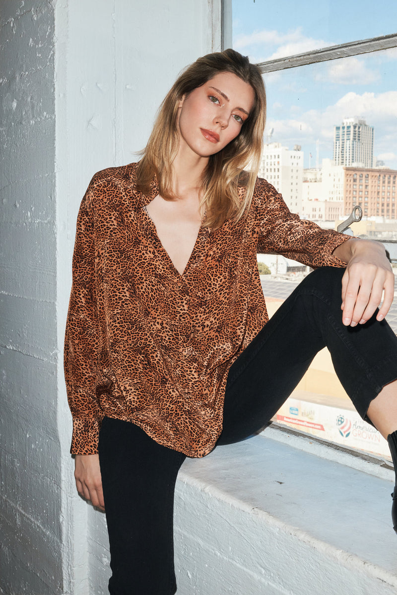 A woman is sitting on a windowsill wearing a Catherine Gee Dakota Blouse in a clean leopard print features a deep V-neck and long sleeves, paired with black pants. The background shows an urban cityscape with buildings and a blue sky visible through the window.