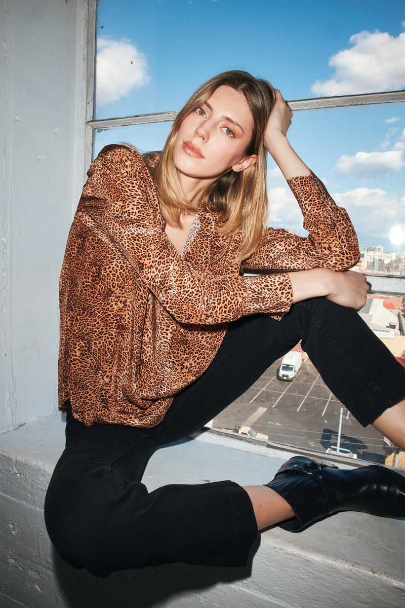 A woman is sitting on a windowsill wearing a Catherine Gee Dakota Blouse - clean leopard. The blouse is loose-fitting with long sleeves paired with black pants and black ankle boots. The background shows a clear blue sky with some clouds and a view of a parking lot and buildings outside the window.