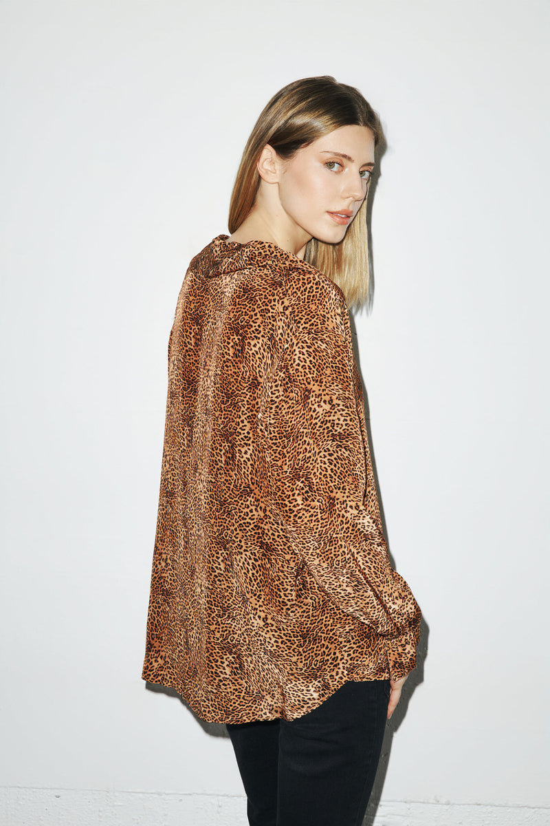 A woman is wearing a Catherine Gee Dakota Blouse in a clean leopard print features a loose, flowing fit with long sleeves and a high neckline. She is standing sideways, showing the side and back of the blouse. The background is plain white, highlighting the detailed leopard pattern of the blouse.