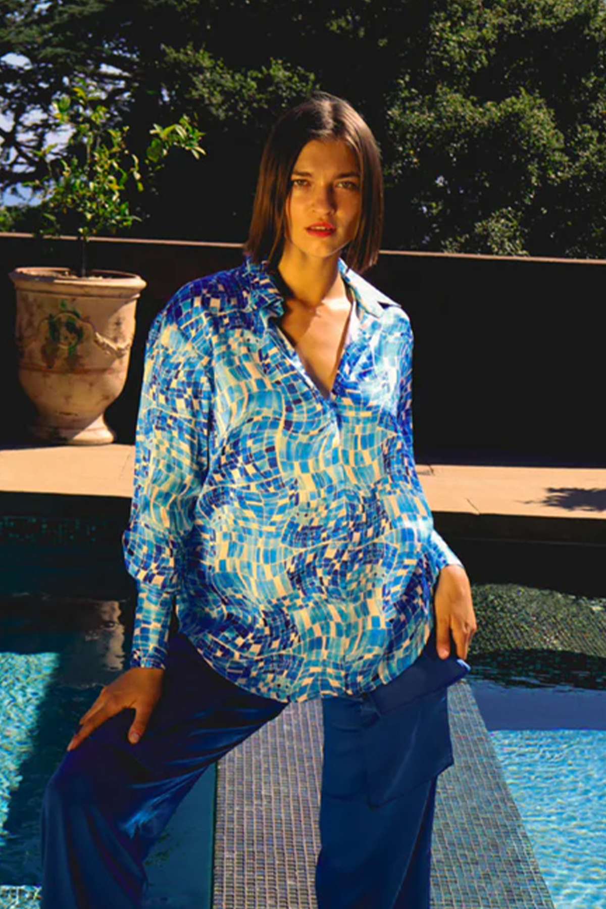 A woman standing by a pool, wearing a Catherine Gee Daria French Cuff Silk Pool Mosaic blouse paired with dark blue pants features a vibrant mosaic pattern in shades of blue and white, resembling the tiles of a swimming pool. The background includes a large potted plant and lush greenery, suggesting an outdoor setting, and highlighting the intricate design and luxurious fabric of the blouse, making it a standout piece for fashion enthusiasts.