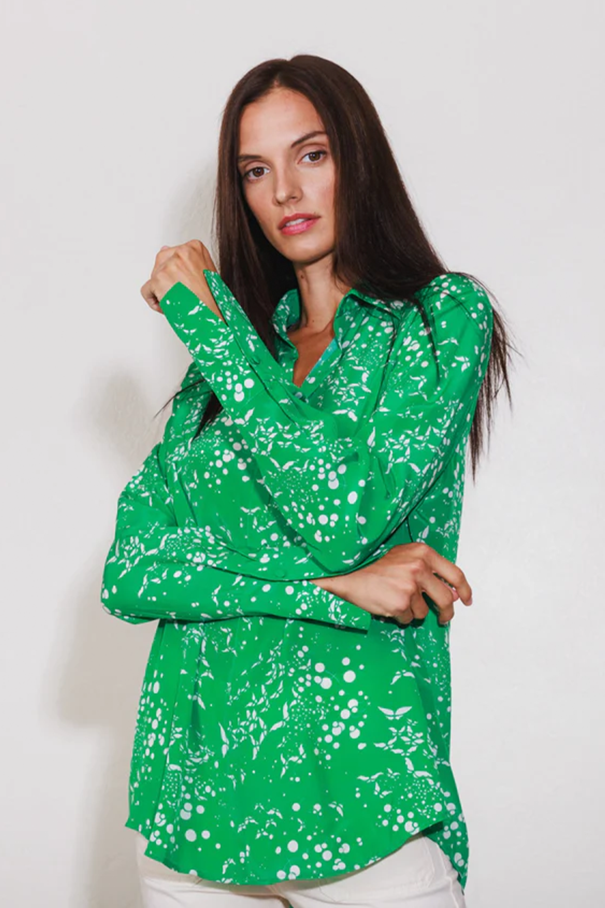 A woman wearing a Catherine Gee Daria French Cuff Silk - Celestial Butterfly blouse, features green with a white abstract pattern resembling butterflies and celestial elements. She is posing with one arm crossed and the other hand touching the opposite shoulder on a plain white background, making the blouse stand out prominently.