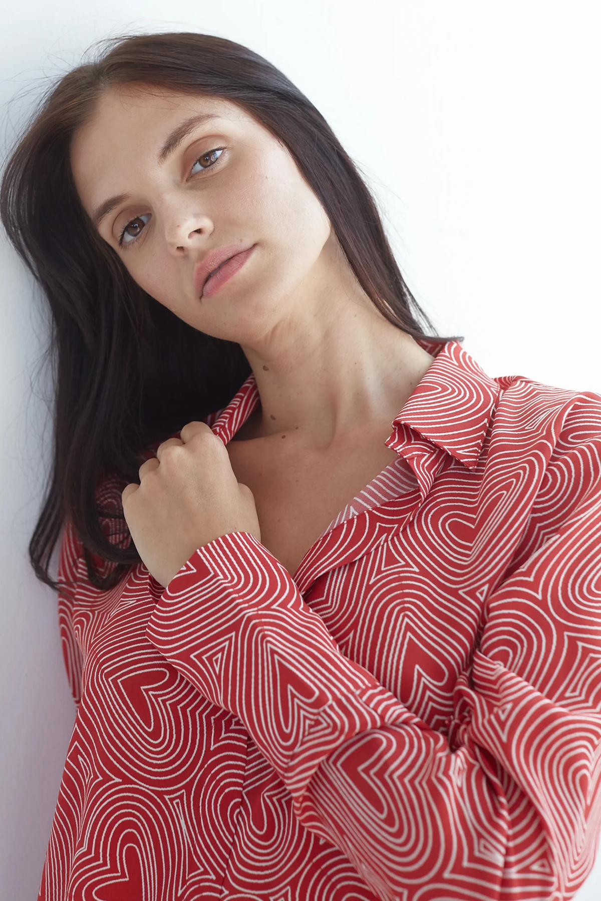 A woman wearing a Catherine Gee Daria French Cuff Silk Blouse - Bowie Heart. The blouse is red with a white heart pattern, featuring a classic collar and long sleeves with French cuffs. She is standing against a white background, holding the collar of the blouse highlighting the blouse's design.