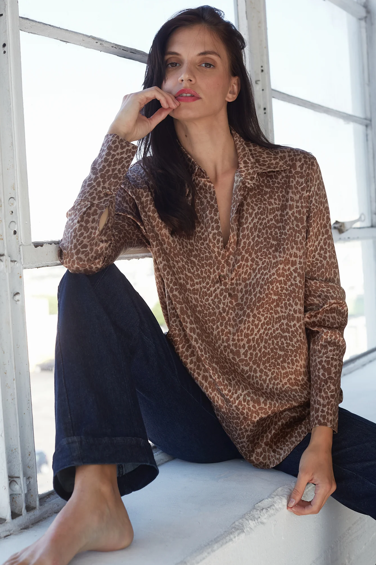 A woman sitting on a windowsill wearing a Catherine Gee Daria French Cuff Silk-Electric Leopard blouse features a leopard print pattern in shades of brown and beige paired with dark blue jeans and is barefoot. The setting appears to be indoors with natural light coming through the window, creating a relaxed and casual atmosphere.
