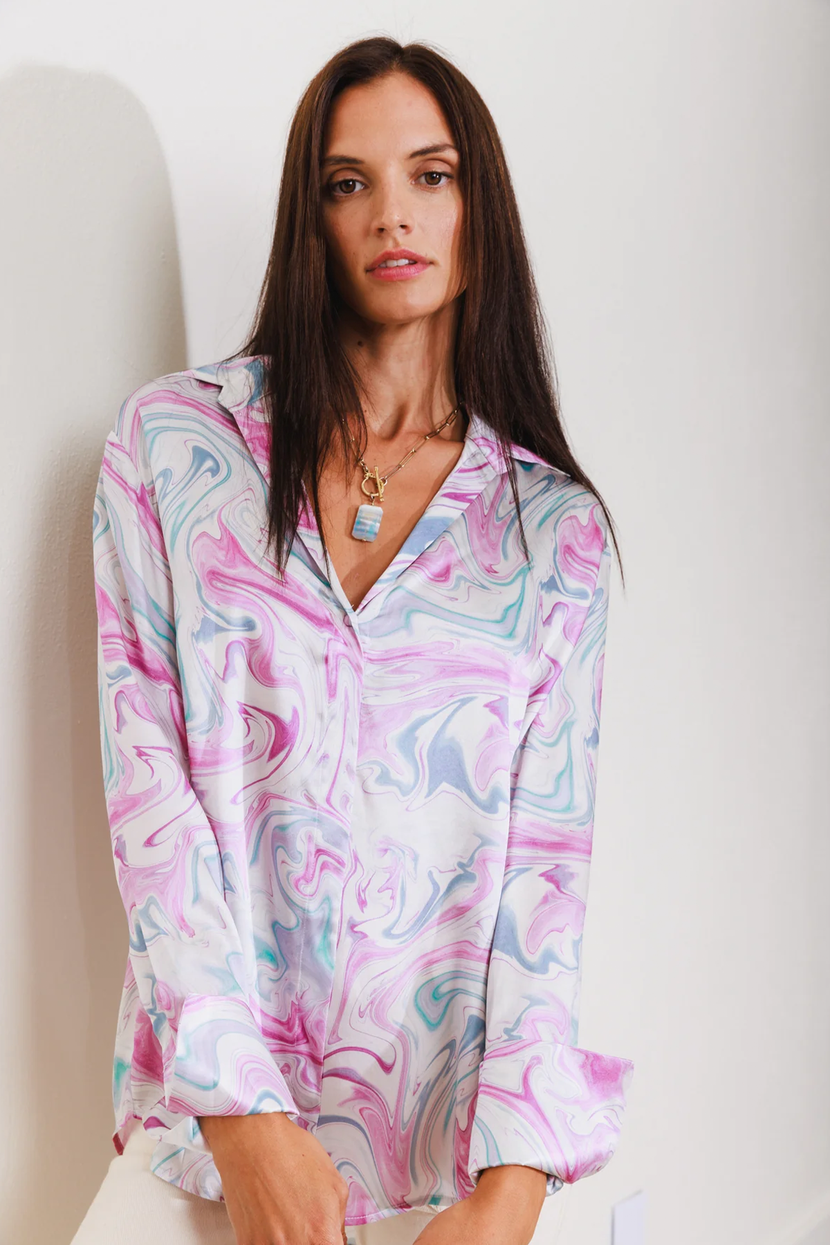 A woman wearing a Catherine Gee Daria French Cuff Silk-Girl Boy Swirl blouse features a colorful, swirling pattern with shades of pink, blue, and white. She is also wearing a necklace with a rectangular pendant on a plain white background.