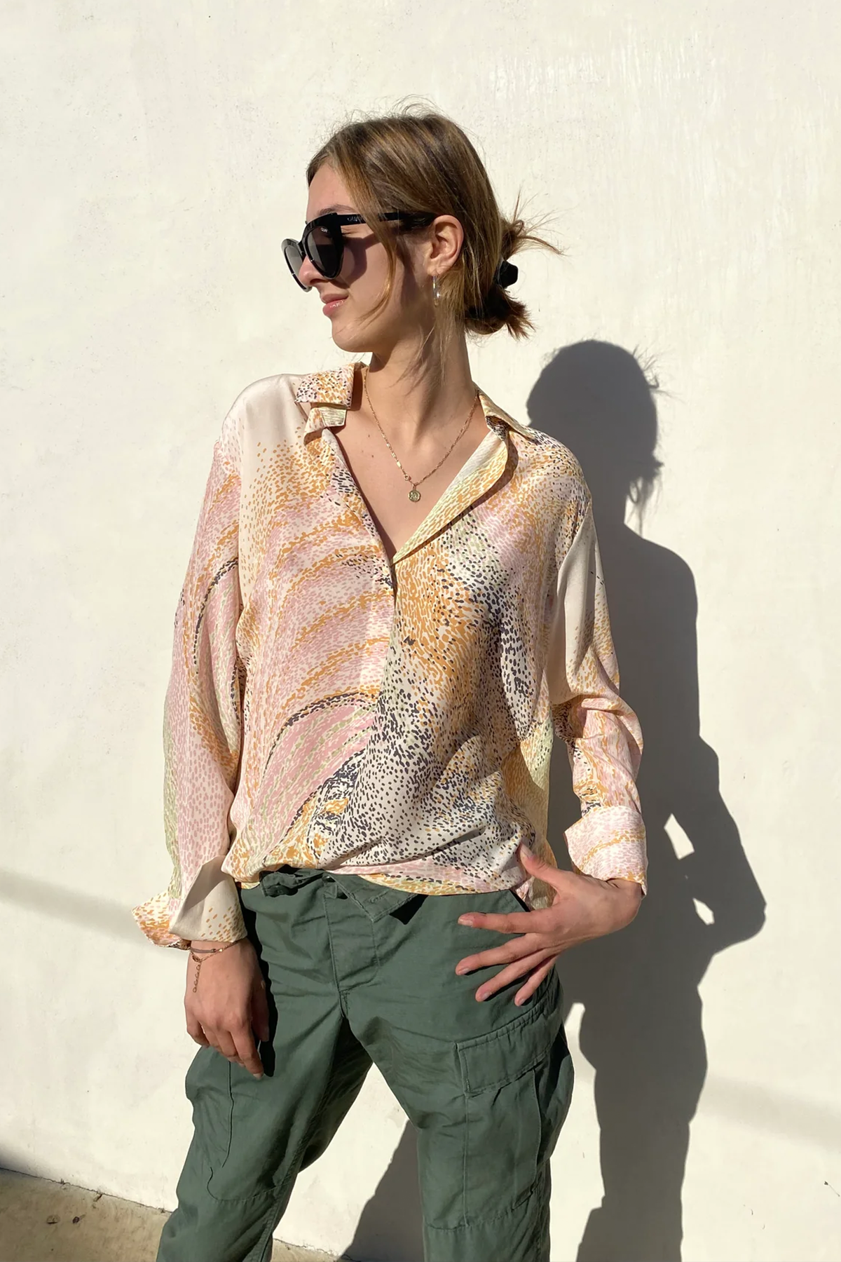 A woman wearing a Catherine Gee Daria French Cuff Silk Orbit blouse features a vibrant, abstract pattern with shades of pink, orange, and yellow, creating a dynamic and eye-catching design. It has a loose, flowing fit with a V-neckline and French cuffs on the sleeves paired with green cargo pants, adding a casual yet stylish contrast to the elegant blouse. The background is a plain, light-colored wall with a shadow cast on it, highlighting the blouse's colorful and unique print.