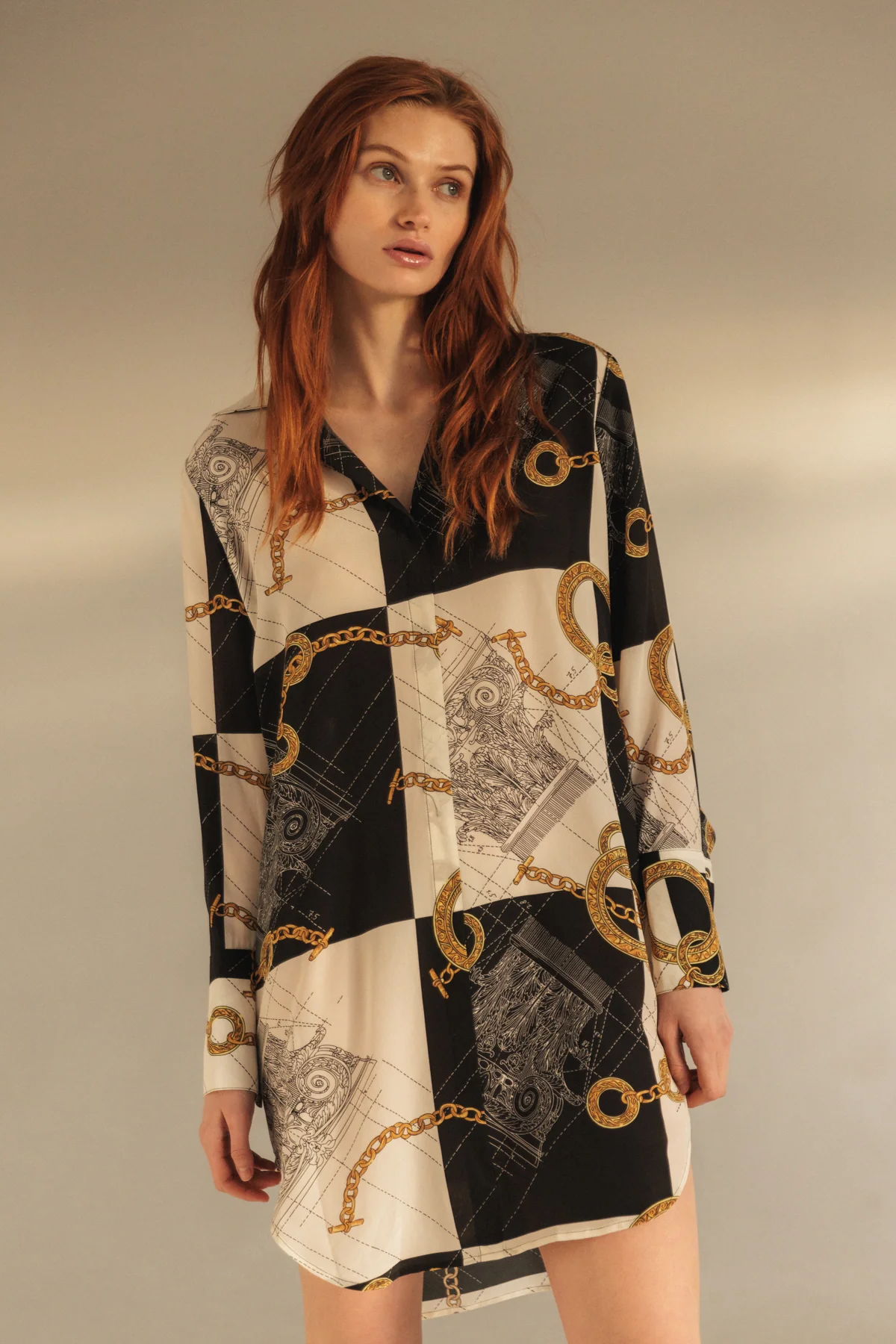 A woman wearing the Catherine Gee Daria Silk Shirt Athens Dress. This elegant dress features a black-and-white color scheme with a complex gold chain and architectural print patterns. It has a loose, shirt-like design with a pointed collar and long sleeves, giving it a sophisticated and timeless look. The silk fabric adds a luxurious touch, enhancing its smooth and elegant appearance.