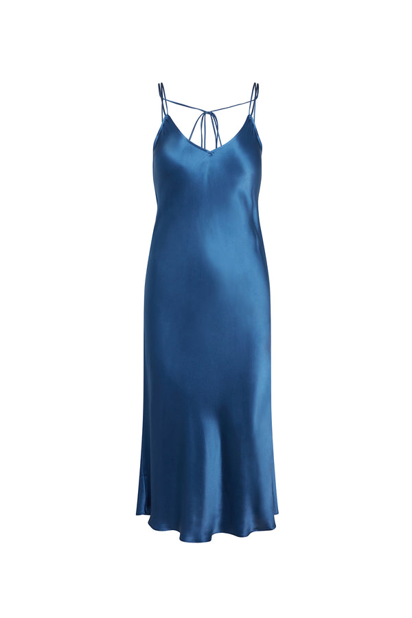 Catherine Gee Emma Cannes Blue Slip Dress. This elegant dress features a rich blue color and a silky, smooth texture, and has a V-neckline with thin spaghetti straps that tie at the back, creating a delicate and sophisticated look. The dress falls gracefully to a midi length, with a slight flare at the hem, making it perfect for both casual and formal occasions. The luxurious fabric and timeless design make this dress a standout piece.