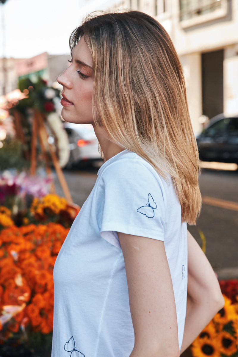 A woman standing near a flower market, with vibrant flowers in the background wearing a white Catherine Gee Embroidered Cotton Butterfly T-Shirt. This elegant shirt features small, delicate butterfly embroidery on the sleeve and side. The t-shirt’s minimalist design is balanced with whimsical details, making it a perfect blend of casual and chic for everyday wear.