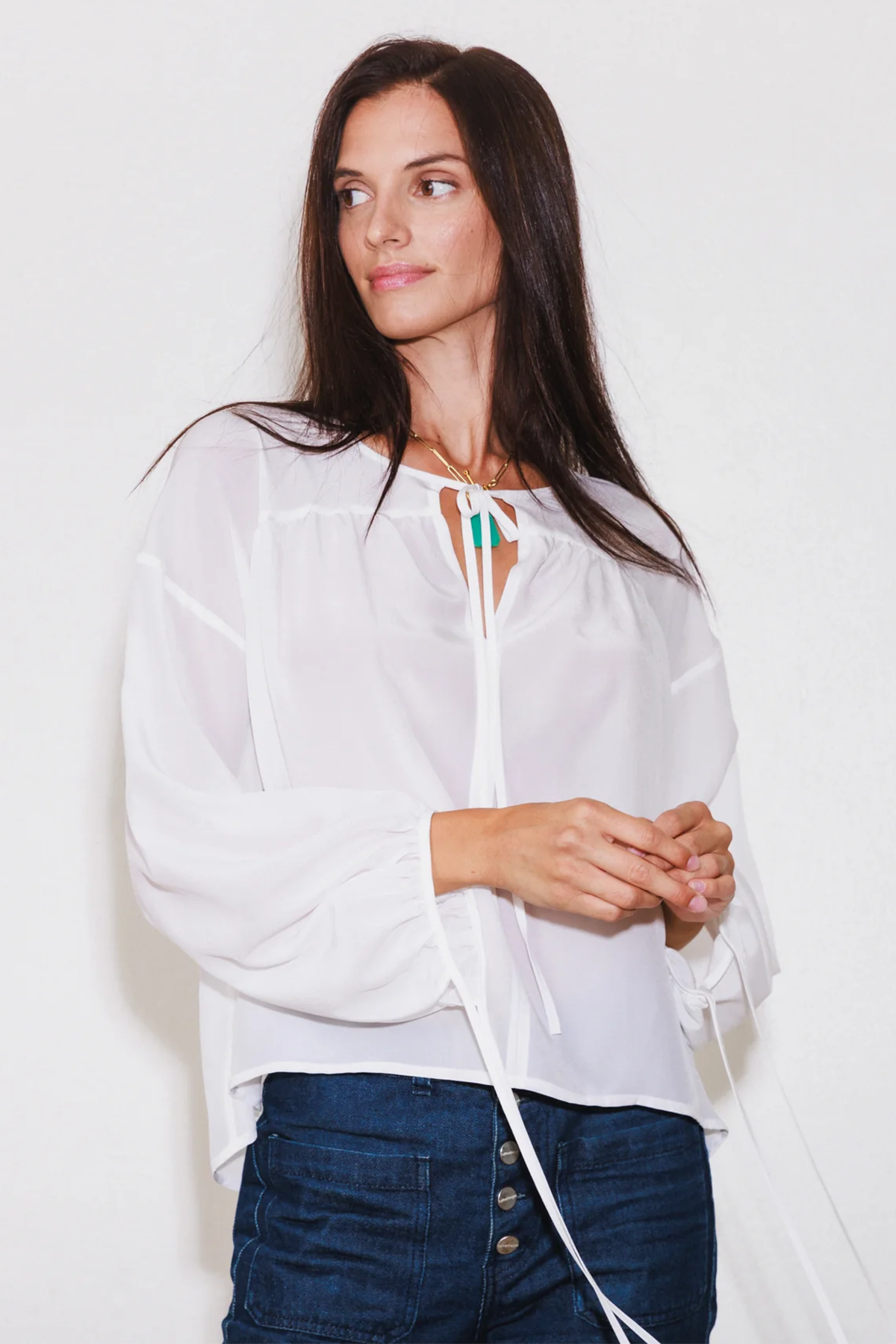A woman wearing the Catherine Gee Frances White Blouse. This semi-sheer blouse features a loose, flowing design with long sleeves and a tie detail at the neckline paired with dark blue jeans that have multiple front buttons. The blouse's light, airy fabric, and elegant design make it suitable for various occasions, blending casual comfort with a touch of sophistication.