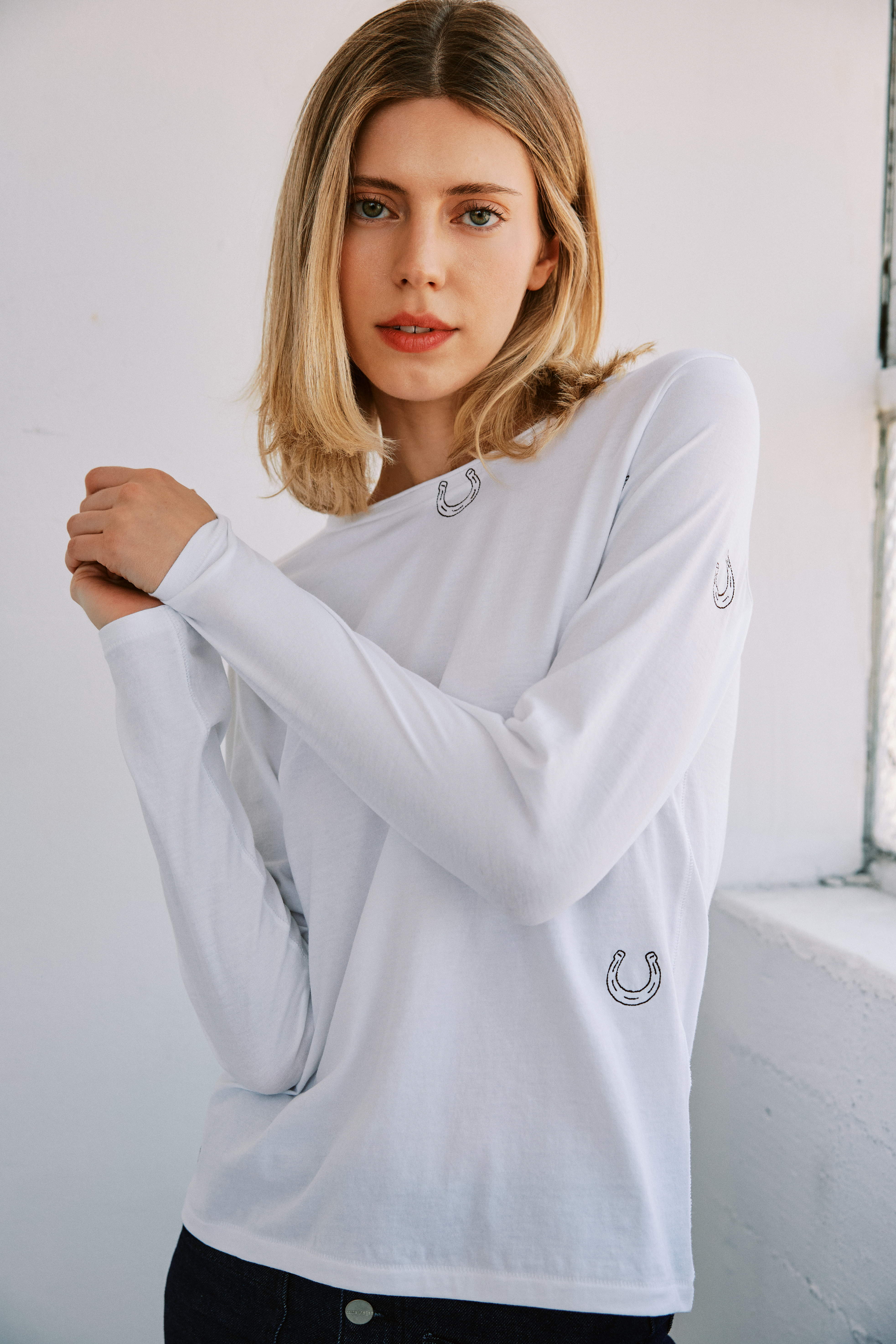 A woman standing in a well-lit room with a white wall and window in the background wearing a Catherine Gee Embroidered Cotton Long Sleeve white Horseshoe T-Shirt features small embroidered horseshoe designs on the front and sleeves paired with dark jeans. The casual fit and unique horseshoe embroidery make this t-shirt a stylish and comfortable choice for everyday wear, enhancing the shirt's clean and minimalist look. 