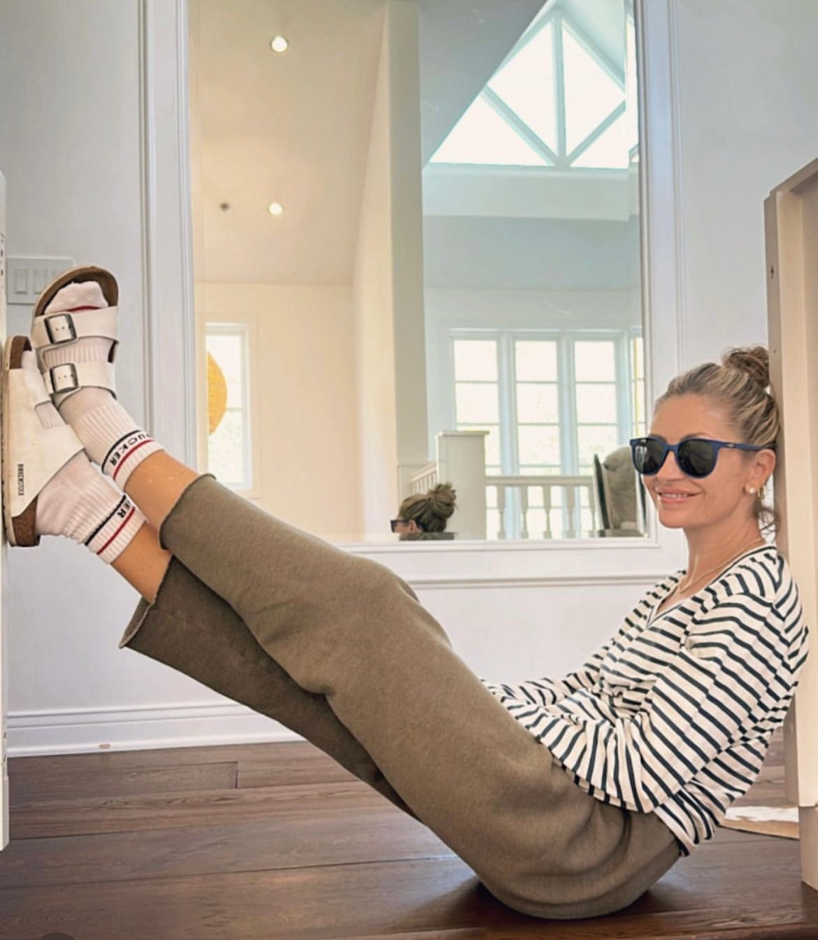 A woman sitting on the floor in a relaxed pose, wearing a Catherine Gee Jen Relaxed Longsleeve - Navy Stripe Tee paired with brown pants, white socks, and white sandals. The tee features classic navy and white horizontal stripes, a V-neckline, and long sleeves. The background features a large mirror reflecting a bright room with high ceilings and large windows, adding a sense of spaciousness to the scene.