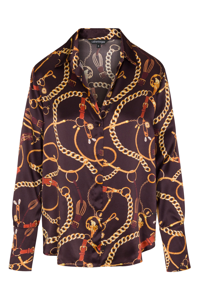 Catherine Gee Daria French Cuff Silk Blouse Belgian Chain with a dark base color, featuring an intricate pattern of gold chains, belts, and equestrian-themed elements. The blouse has a classic collar and French cuffs, giving it a sophisticated and elegant appearance. The design is luxurious and eye-catching, making it a statement piece suitable for formal or upscale casual occasions.