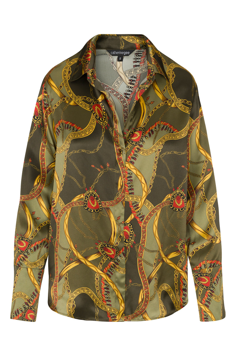 Catherine Gee Daria French Cuff Silk Olive Equestrian blouse features a luxurious silk fabric with an olive green base color. The design is adorned with an intricate equestrian-themed print, including elements such as chains and bridles in gold, red, and black hues. The blouse has a classic collar and button-down front, with long sleeves that end in French cuffs. The overall aesthetic is elegant and sophisticated, making it a standout piece for a refined wardrobe.