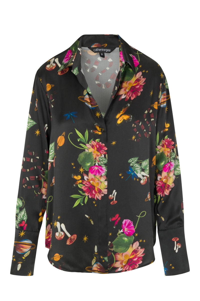 Catherine Gee Daria French Cuff Silk Serpentine blouse features a black background adorned with a vibrant and eclectic print featuring various elements such as flowers, mushrooms, snakes, butterflies, and planets. The design is colorful and intricate, making the blouse visually striking and unique and has a classic collar and button-down front, with long sleeves ending in French cuffs, adding a touch of elegance to the whimsical pattern.