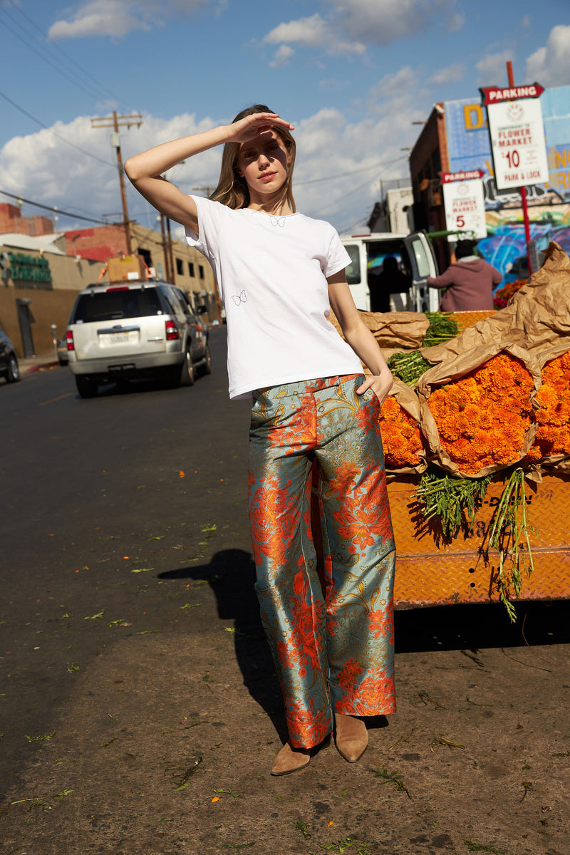 A woman standing next to a cart filled with vibrant orange flowers, wearing the Catherine Gee Jacquard Stella Blue/Orange Pants. These high-waisted pants feature a wide-leg cut and an intricate jacquard pattern in striking blue and orange hues paired with a simple white T-shirt. The urban setting, complete with parked cars and market signage, adds to the lively and fashionable atmosphere.