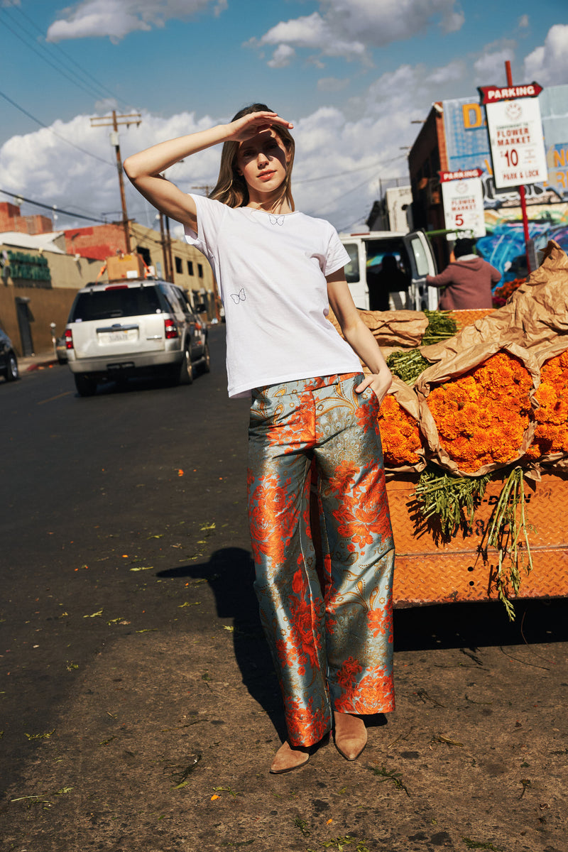 A woman standing on a street, wearing a white Catherine Gee Embroidered Cotton Butterfly T-Shirt paired with metallic, floral-patterned pants features delicate butterfly embroidery, with one butterfly near the neckline and another on the left side. She is posing with one hand on her head and the other resting on her pocket. The urban background includes parked cars and buildings under a partly cloudy sky, adding charm and vibrancy to the shirt.