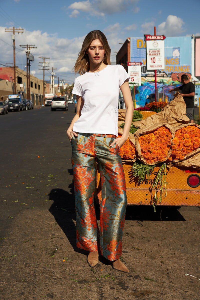 A woman standing on a street wearing Catherine Gee Jacquard Stella Blue/Orange pants features a vibrant, intricate pattern with blue and orange hues that create a striking visual contrast. She pairs the pants with a simple white T-shirt and brown shoes. In the background, there's a cart filled with orange flowers, adding to the colorful and lively atmosphere of the scene.