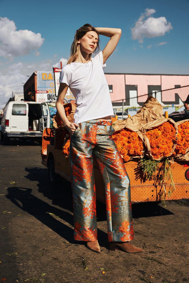 A woman standing in front of a cart filled with vibrant orange marigold flowers wearing a white Catherine Gee Embroidered Cotton Butterfly T-Shirt and colorful, patterned pants features delicate butterfly embroidery, adding a touch of whimsy to the outfit. The background includes a white van and buildings with graffiti, creating a lively urban scene. She is posing with one hand on her hip and the other shielding her eyes from the sun, capturing a candid, stylish moment.