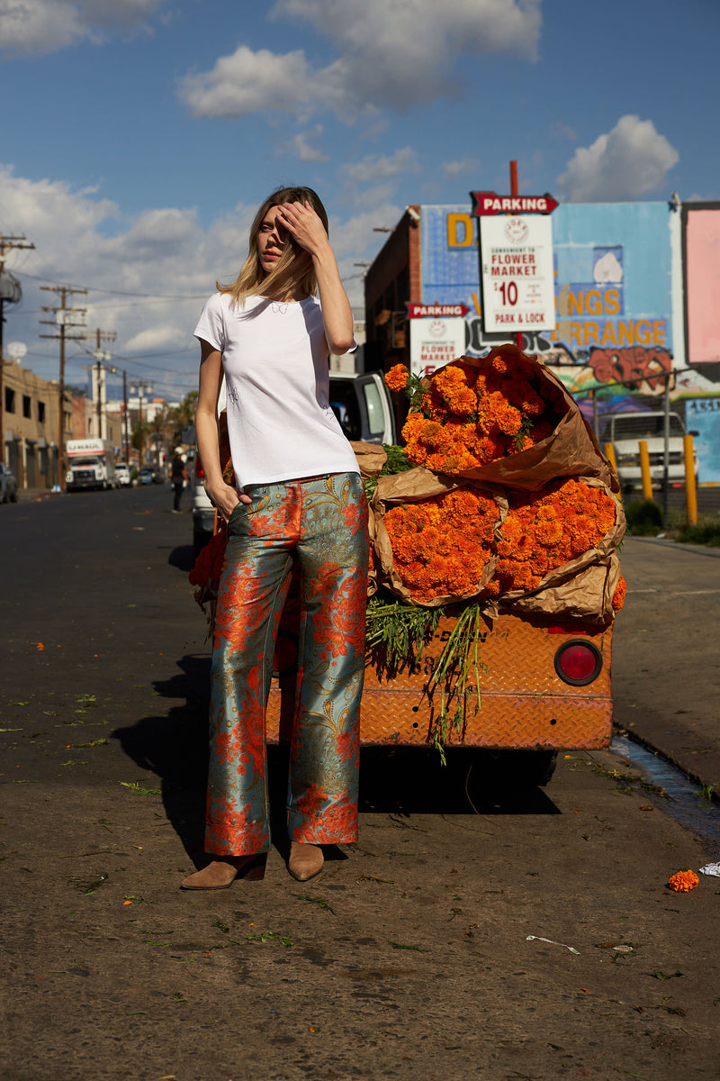 A woman standing in front of a truck filled with vibrant orange marigold flowers wears a white t-shirt and Catherine Gee Jacquard Stella Blue/Orange Pants. These high-waisted pants feature an intricate jacquard pattern with blue and orange floral designs, perfectly complementing the flowers in the truck. The urban street scene in the background adds to the lively and colorful atmosphere.