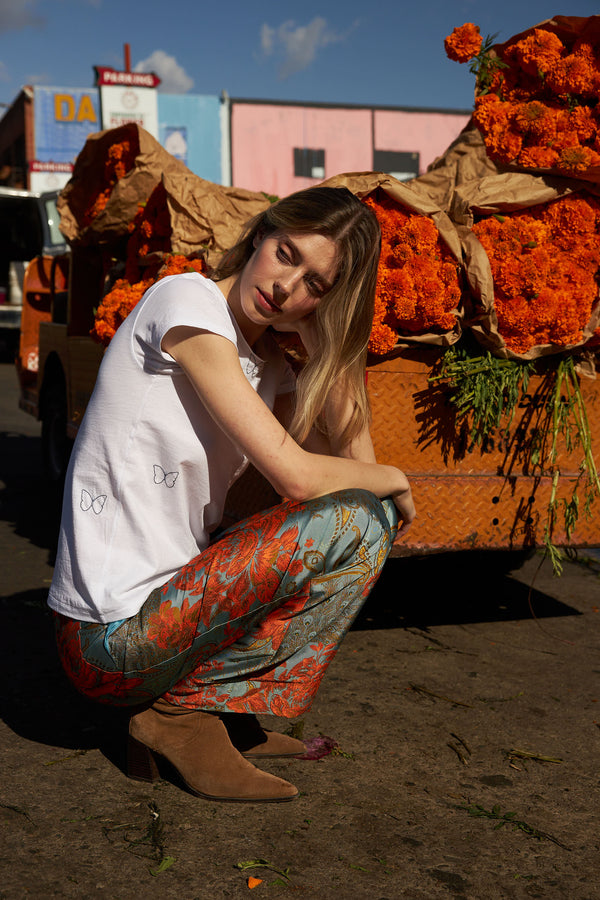 A woman squatting next to a truck loaded with vibrant orange marigold flowers wearing a white t-shirt paired with Catherine Gee Jacquard Stella Blue/Orange Pants, which feature an intricate floral pattern in blue and orange hues. The scene is set outdoors, likely at a market or flower shop, highlighting the bold and colorful contrast between the pants and the marigold flowers. This combination creates a visually striking and stylish look.