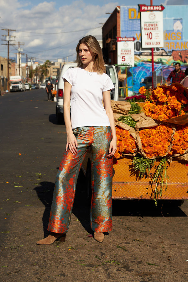 A woman standing on a street wearing the Catherine Gee Jacquard Stella Blue/Orange Pants. These high-waisted, wide-leg pants feature a striking jacquard pattern in blue and orange hues. The outfit is completed with a plain white T-shirt and brown shoes. The backdrop includes a cart filled with vibrant orange flowers and various urban signs and buildings, adding an urban flair to the scene.