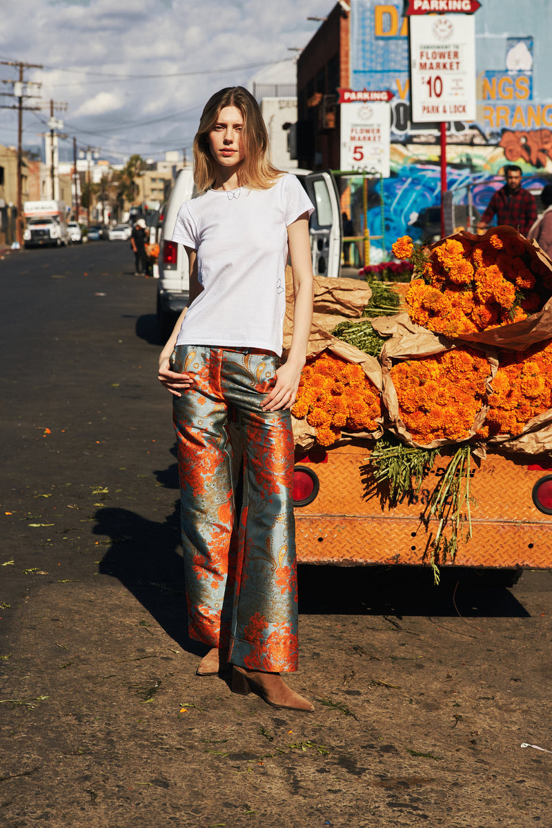 A woman standing in front of a flower market stall, wearing a white Catherine Gee Embroidered Cotton Butterfly T-Shirt features a delicate butterfly embroidery, adding a whimsical touch paired with colorful patterned pants. The background includes a cart filled with bright orange flowers and various signs creating a stylish and vibrant getup that captures the lively atmosphere.