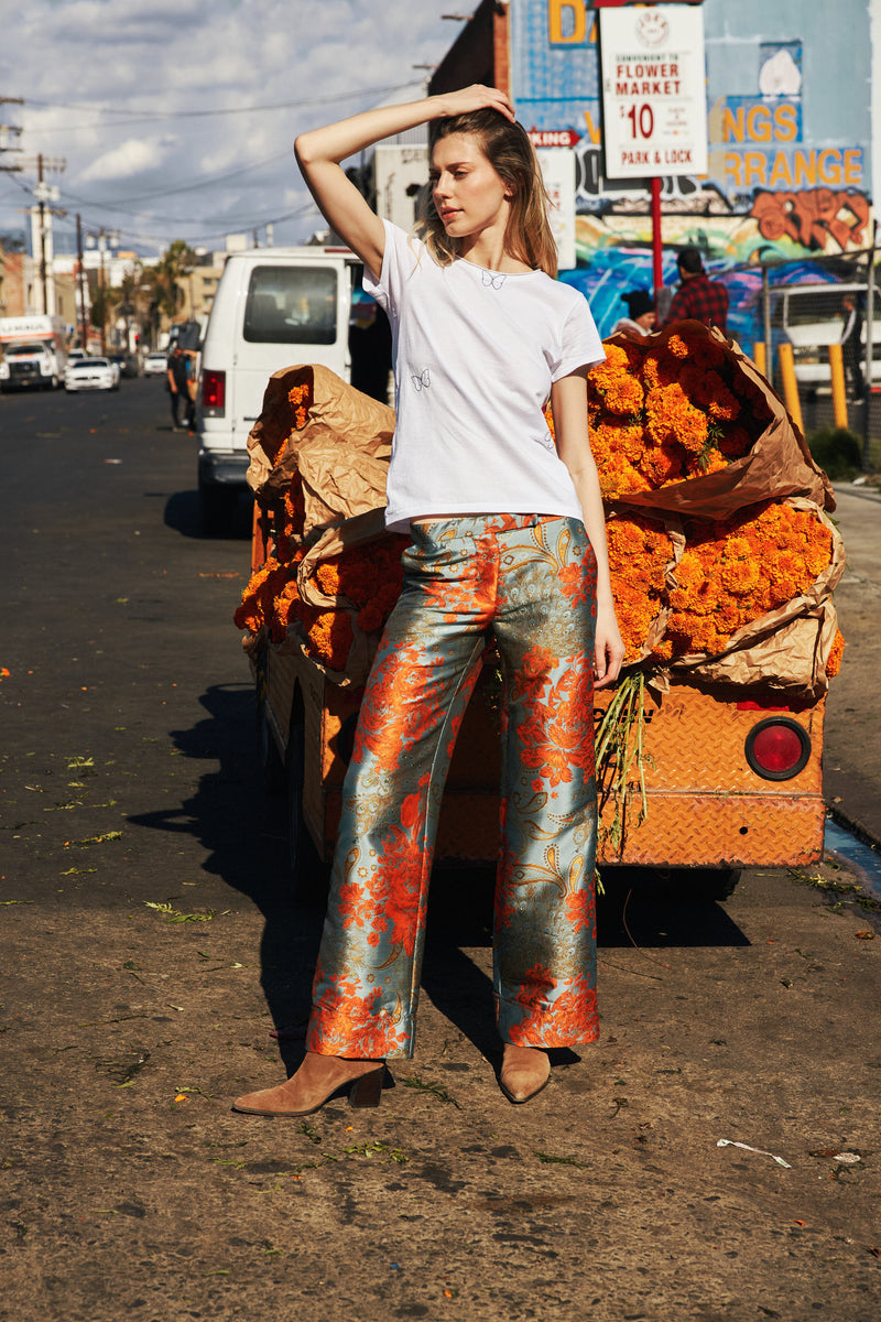 A woman standing in front of a flower market stall, wearing a white Catherine Gee Embroidered Cotton Butterfly T-Shirt features delicate butterfly embroidery on the front, adding a whimsical touch paired with colorful patterned pants that stand out against the vibrant orange marigold flowers in the background. The urban setting, including a van and a street sign, adds a dynamic and lively atmosphere to the scene.