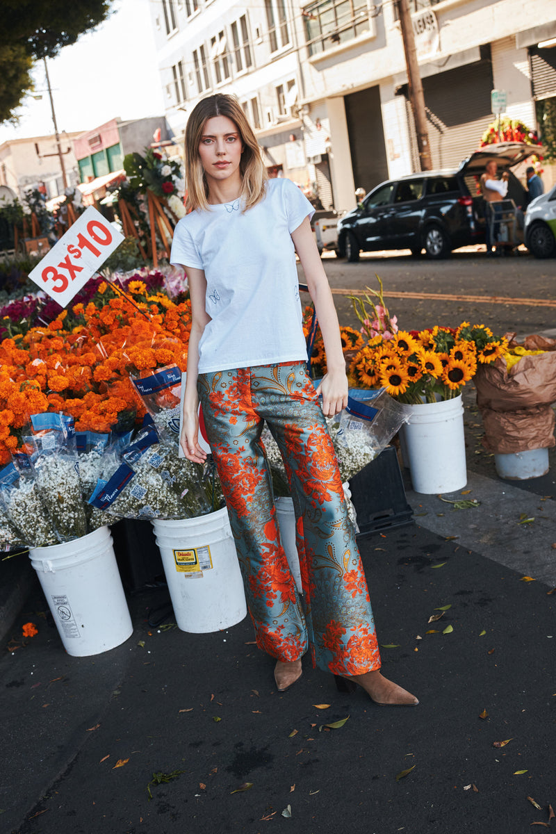 A woman standing in front of a flower stall on a street wearing a white Catherine Gee Embroidered Cotton Butterfly T-Shirt and colorful, patterned pants. The t-shirt features delicate butterfly embroidery, adding a whimsical touch. The background includes a cart filled with vibrant orange and yellow flowers, a sign reading "3x$10," and a lively urban scene. This stylish t-shirt perfectly balances casual elegance with a unique design, making it a standout piece for any wardrobe.