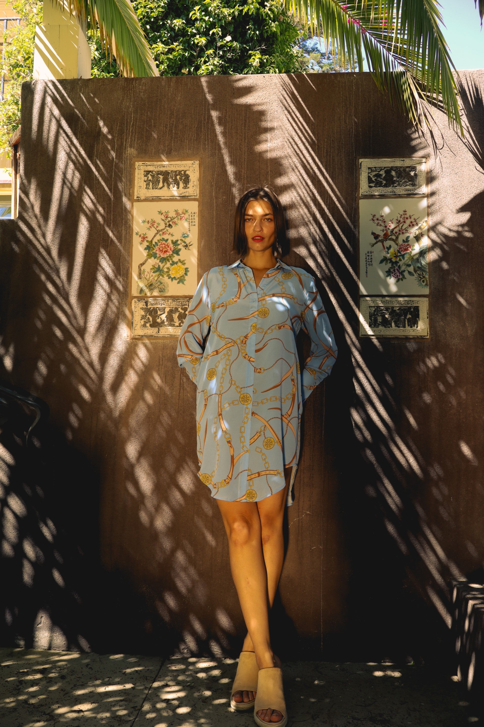 A woman standing against a textured wall adorned with decorative panels featuring floral designs wearing a Catherine Gee Daria Shirt - San Telmo Dress, a loose-fitting, light blue dress with a pattern of golden chains. The dress has long sleeves and exudes a relaxed, casual style paired with beige open-toe sandals. Palm fronds cast shadows, adding depth and elegance to the scene.