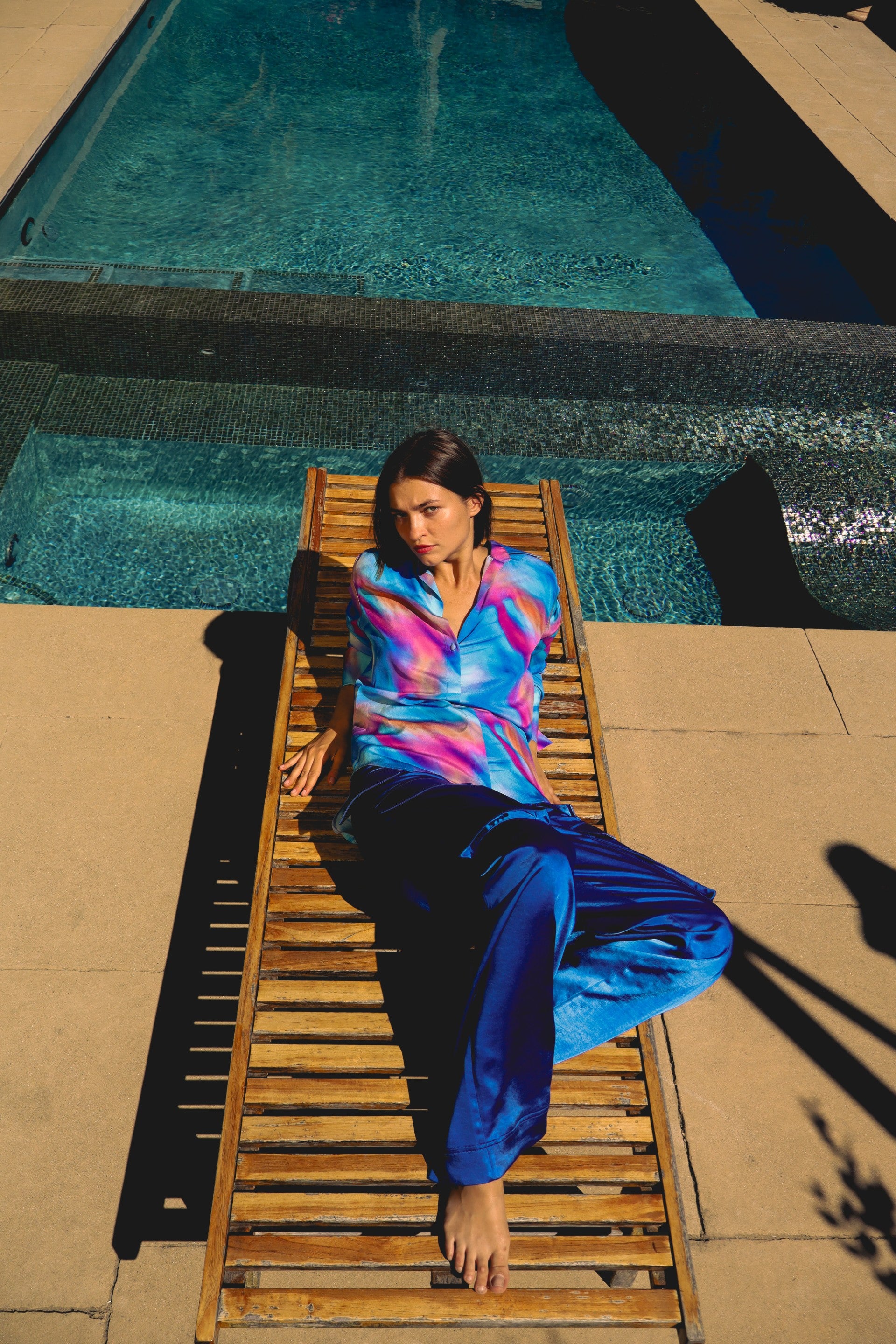 A woman is lounging on a wooden poolside chair wearing Catherine Gee's Cargo Blue Pants and a colorful, tie-dye style shirt. The person is positioned near a clear blue swimming pool, with the water reflecting the sunlight appears to be a sunny day, and the person is relaxing with one leg bent and the other extended on the chair. The background includes the pool and surrounding patio area.